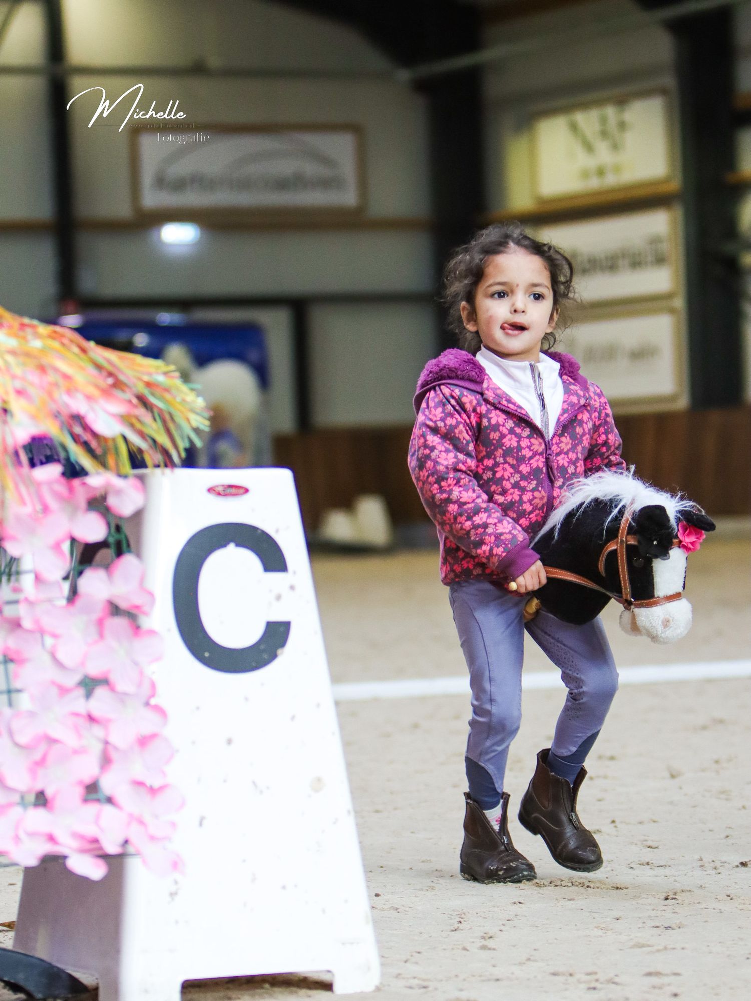 Young girl in pink jacket rides a hobby horse toy in an indoor equestrian arena near a letter C dressage marker.