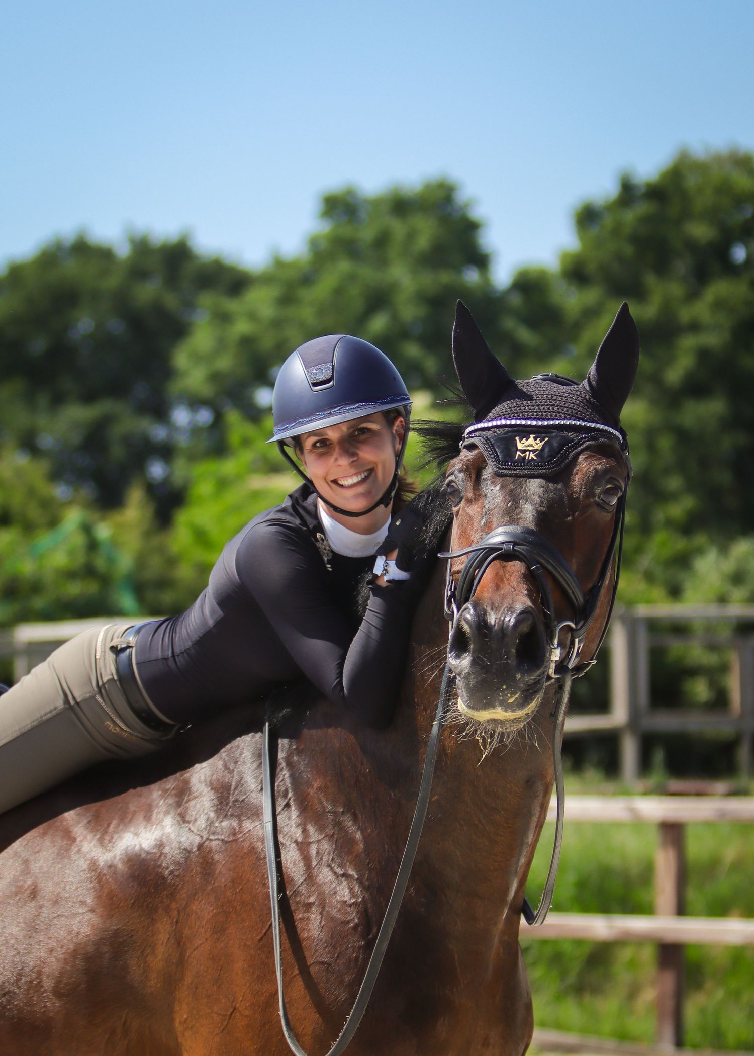 Smiling equestrian rider in navy helmet leaning affectionately on bay horse outdoors on sunny day.