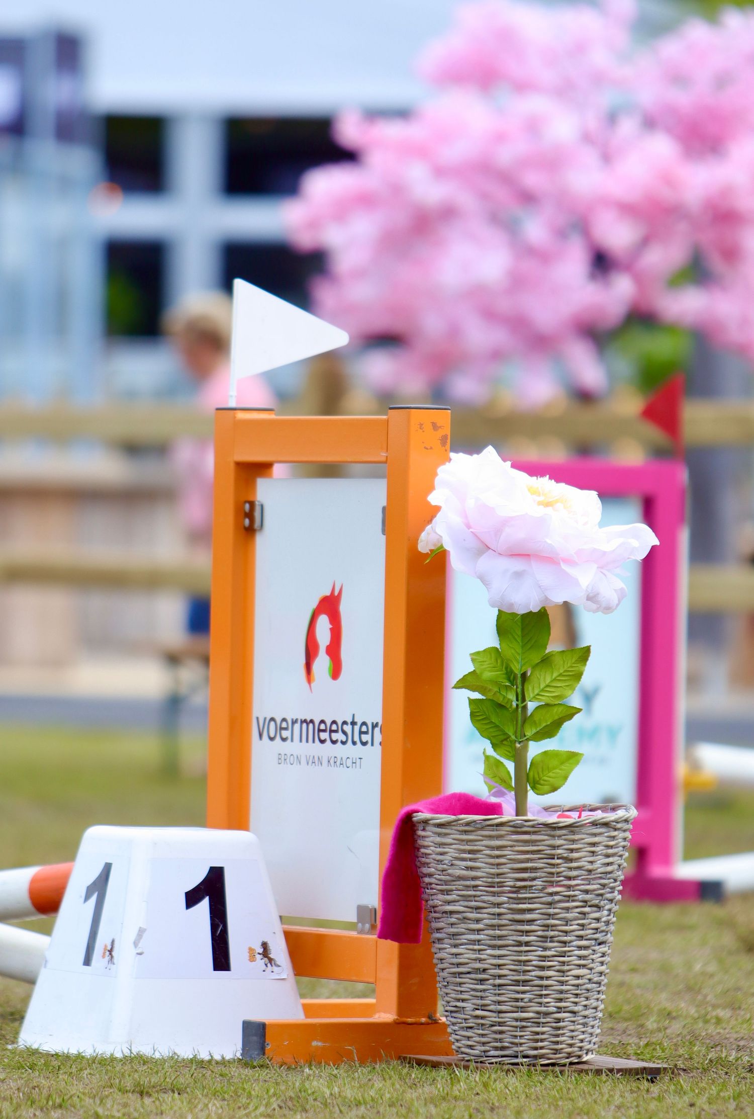 Voermeester branded orange jump standard with number 1 markers and pink rose in wicker pot at equestrian show jumping event.
