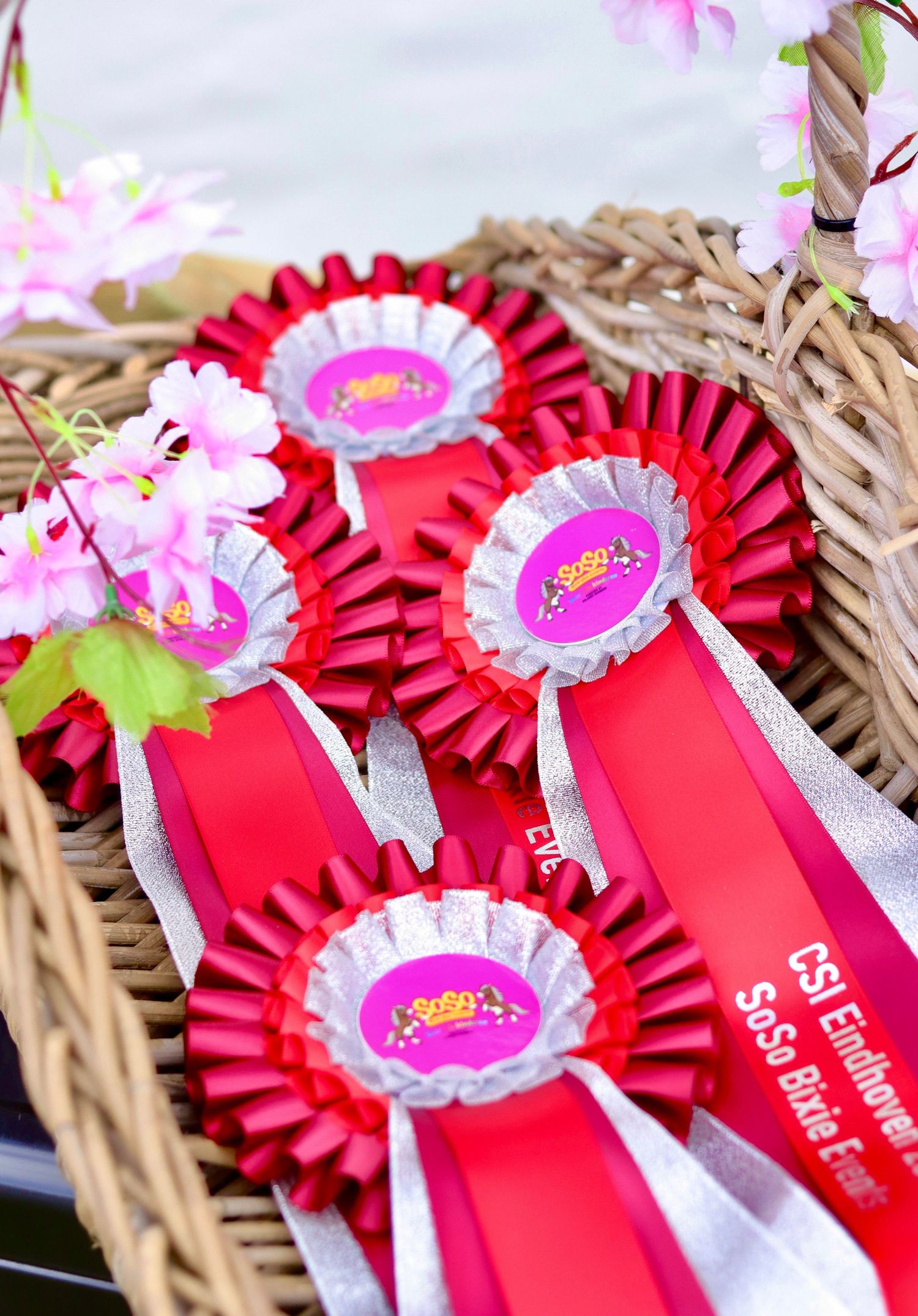 Red and silver rosette award ribbons displayed in a wicker basket with pink flowers at an equestrian event.