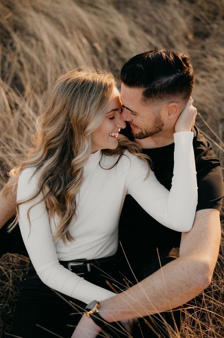 Romantic couple embraces in golden sunlight while sitting in tall dry grass during sunset photoshoot.