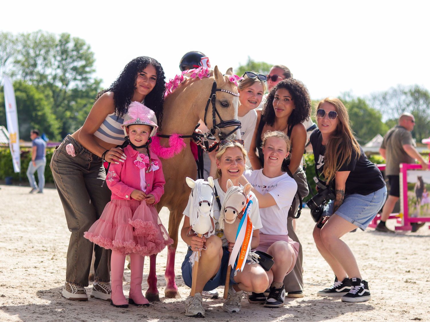Group of girls posing with decorated horses at an outdoor equestrian event on a sunny day.