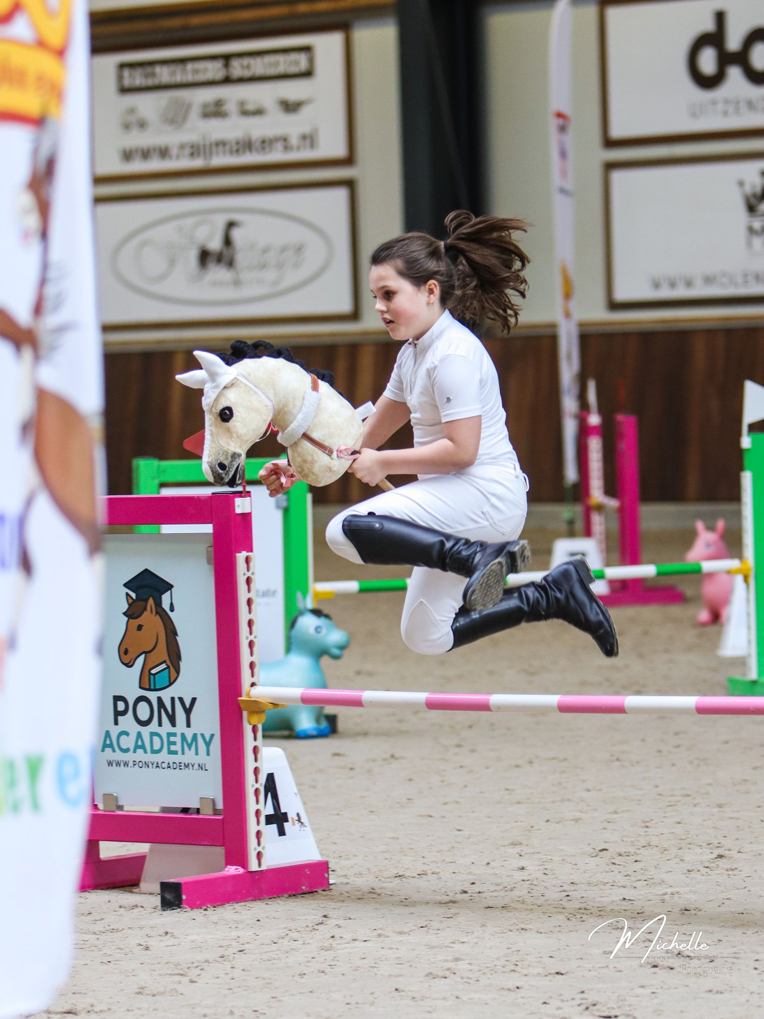Girl jumping over a hurdle with a hobby horse at a Pony Academy competition in an indoor arena.
