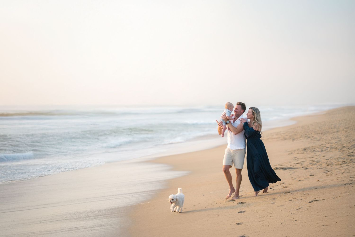 Family walking with a small dog along beach shoreline at sunset in Orange County, California.