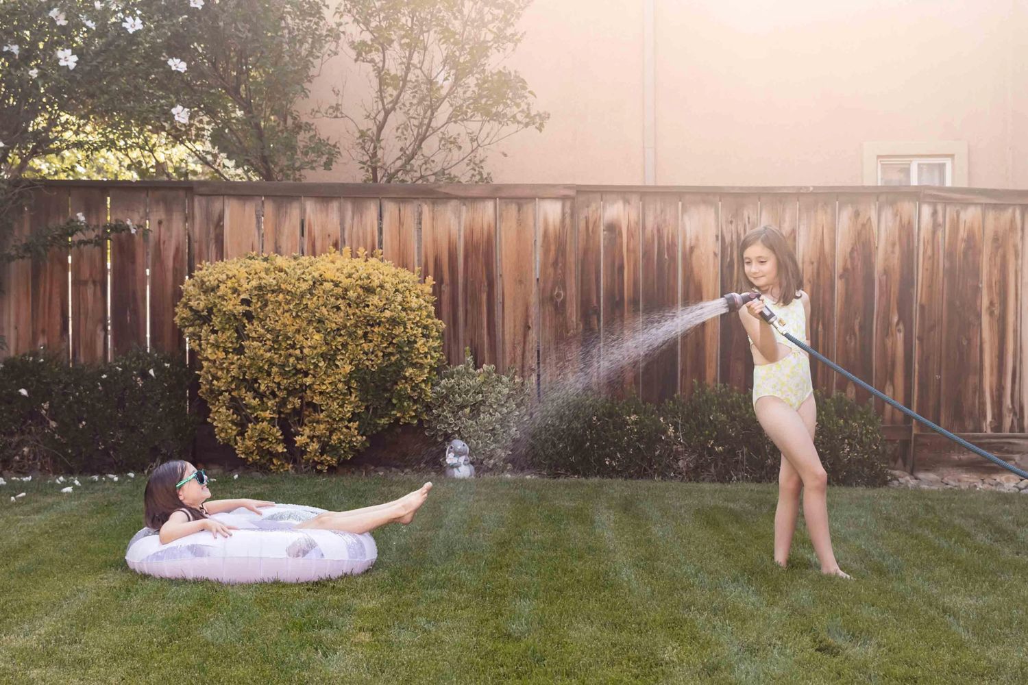 Girls sprays water with hose on sister sitting in a floatie on the lawn