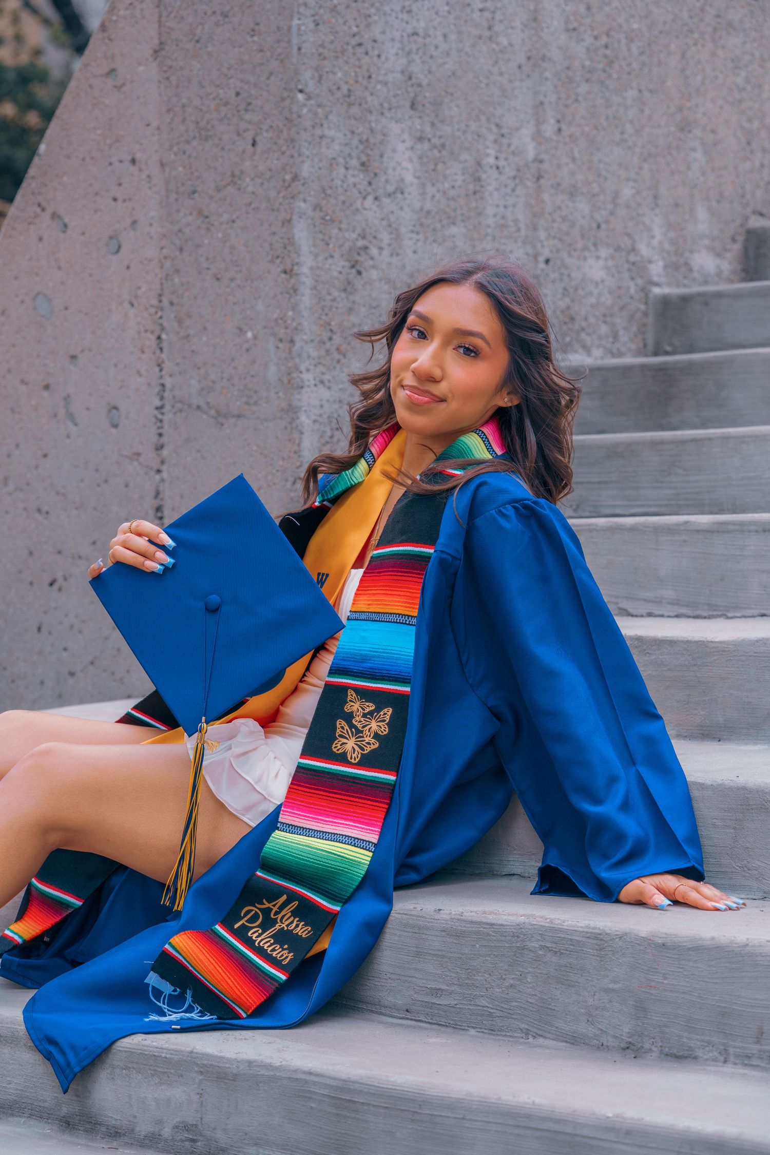 Graduate in blue cap and gown with colorful serape stole poses on concrete steps for graduation photos.