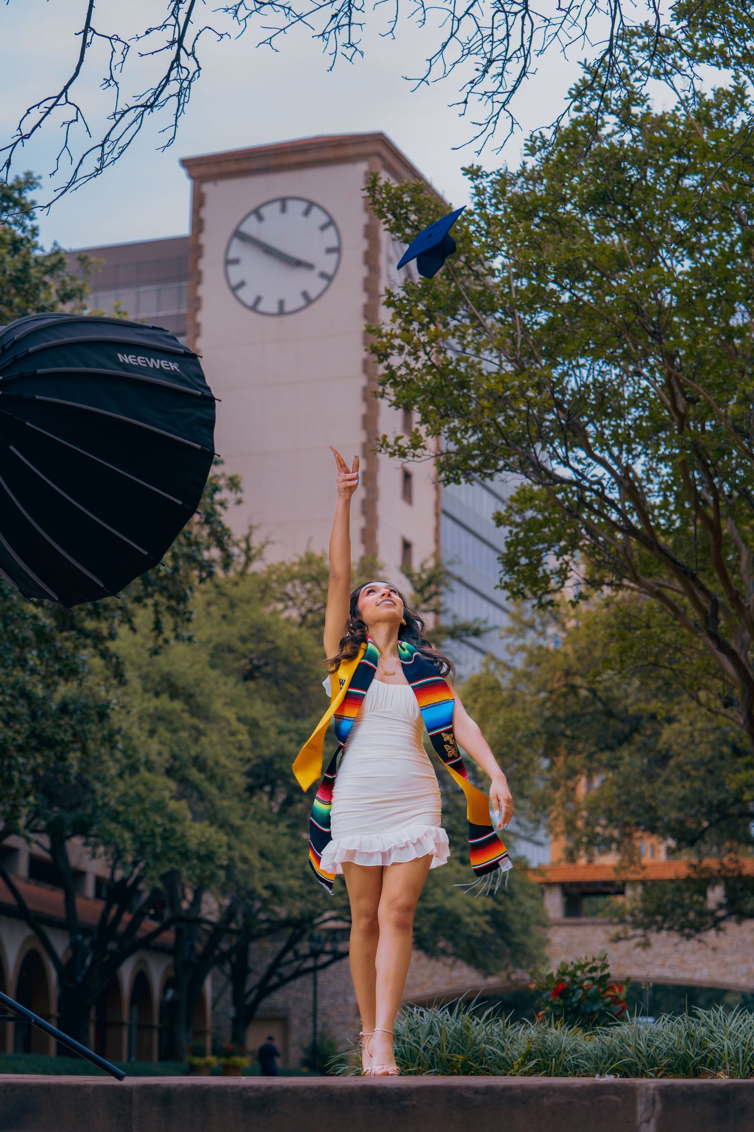 Graduate in white dress and graduation stole jumps joyfully in front of campus clock tower during celebration photos.