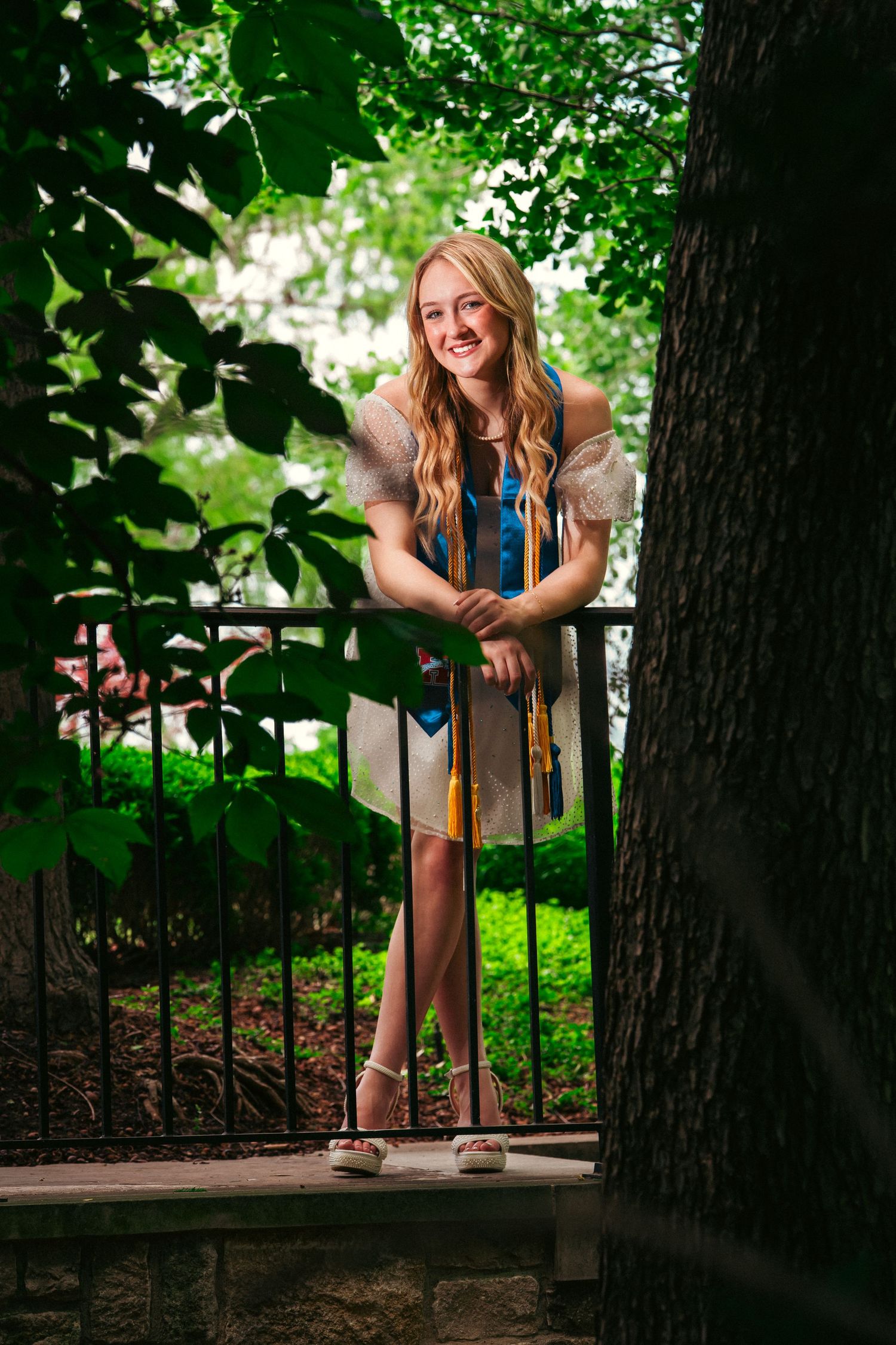 Young woman in blue dress poses playfully near trees and iron fence in outdoor garden setting.