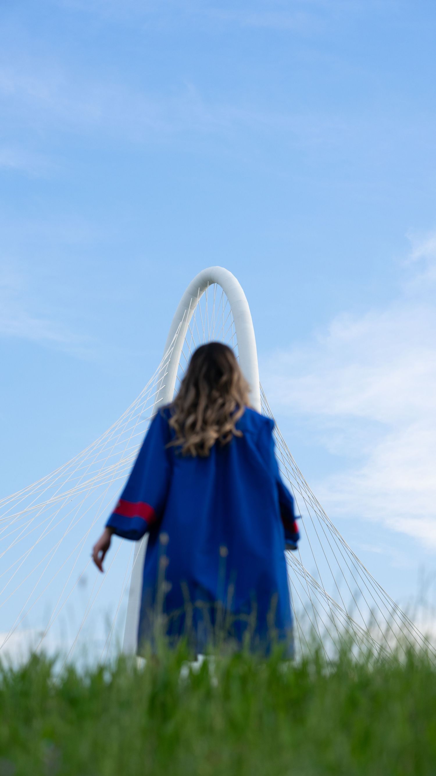 A figure in a blue graduation gown sits on a grassy hill against a bright blue sky.