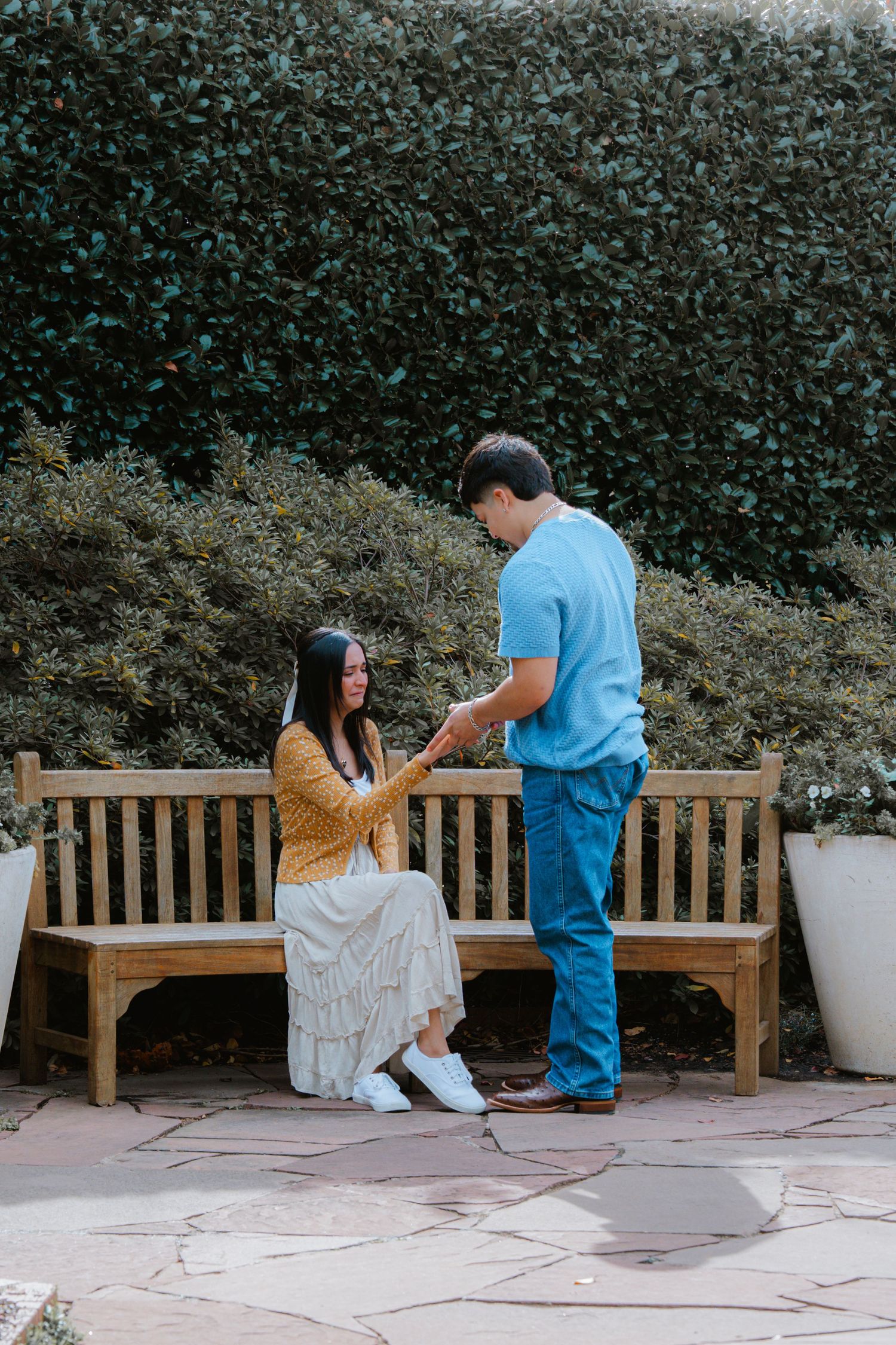 A couple shares a romantic moment on a wooden bench in a garden setting with hedges in the background.