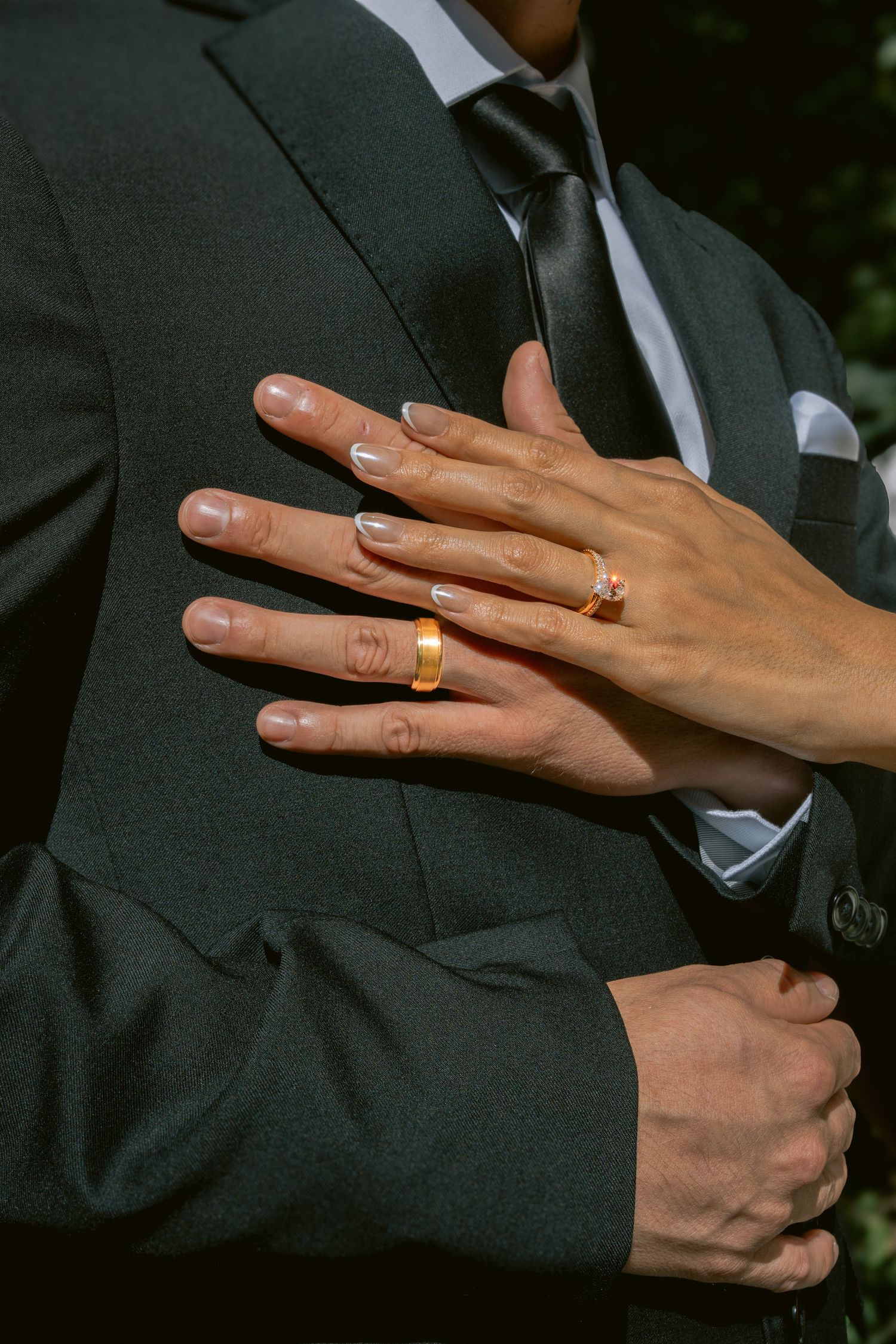 Close-up of newlywed couple's hands showing gold wedding bands, bride's French manicure resting on groom's dark suit.