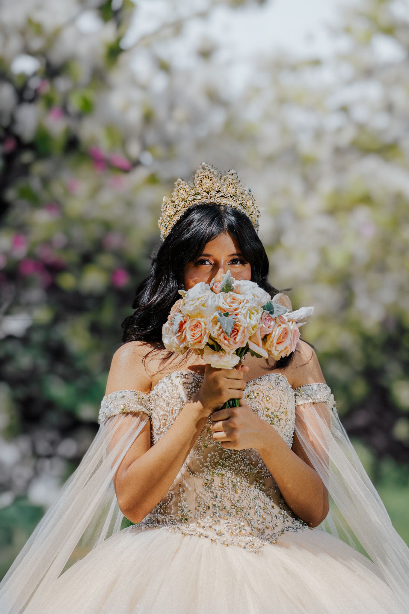 Bride wearing gold crown and embellished ball gown holding pastel rose bouquet near blooming trees.