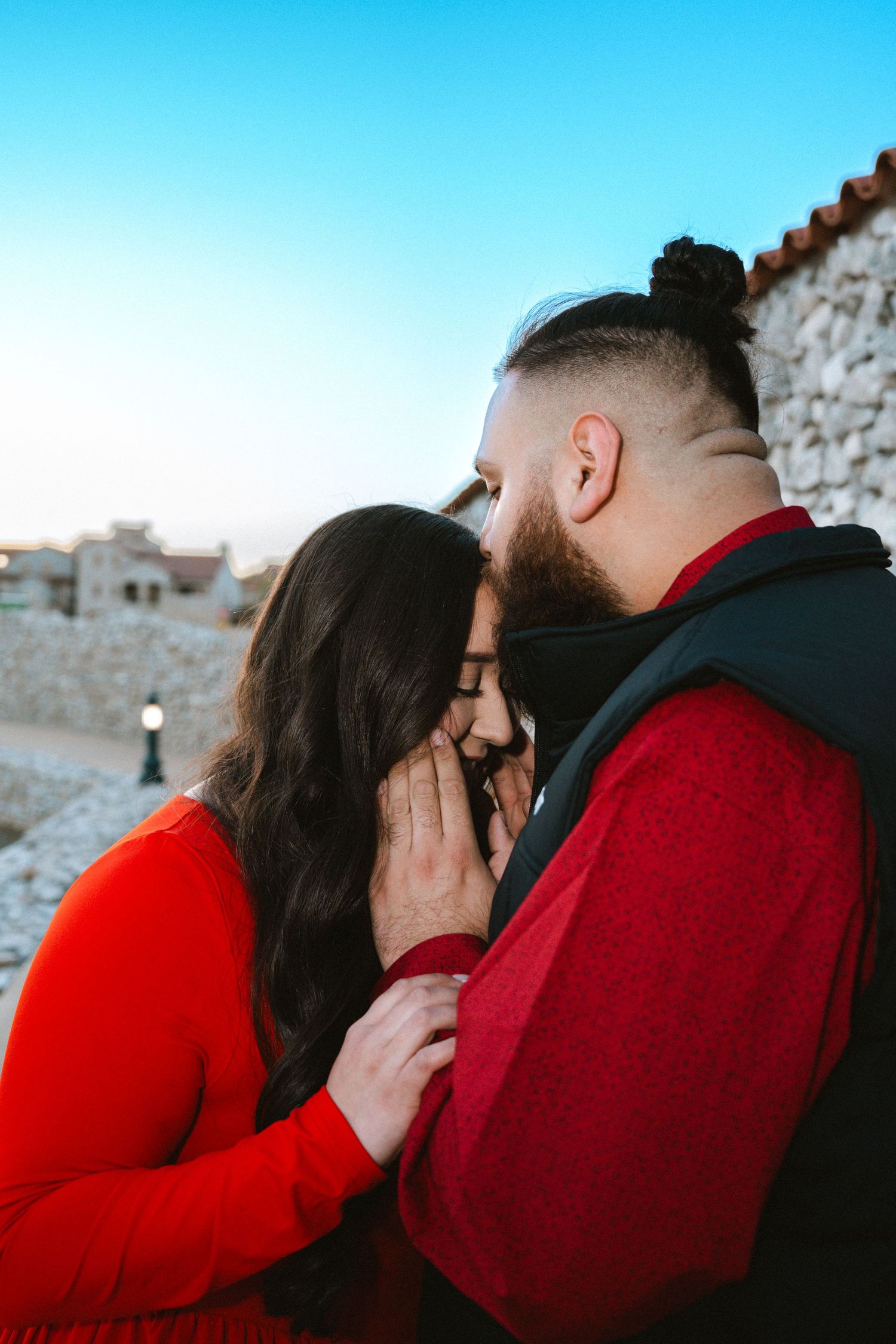A bearded man with a bun hairstyle tenderly holds a woman's face while they share an intimate moment outdoors.