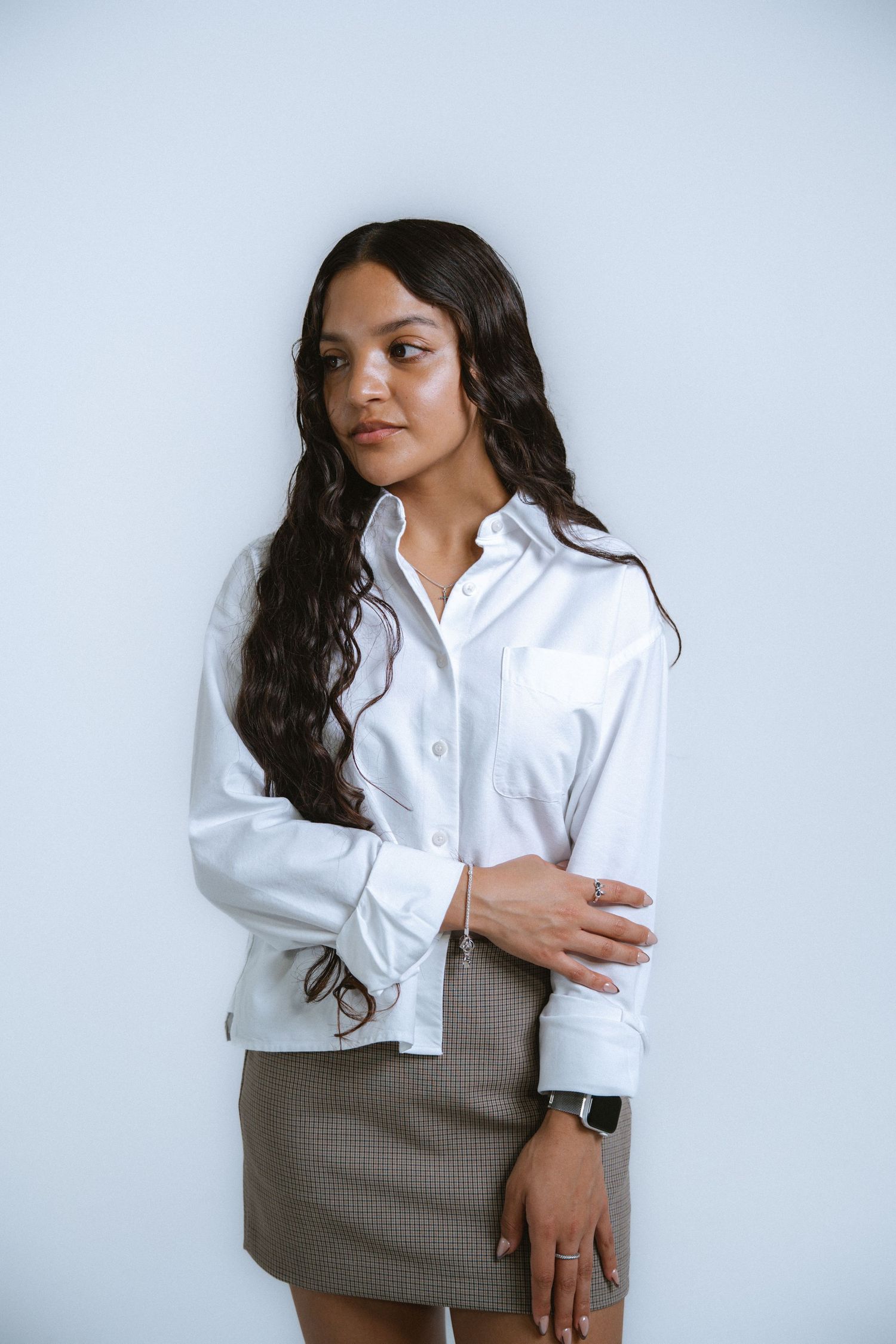 Young woman in white button-up shirt and plaid mini skirt posing against a light gray background.