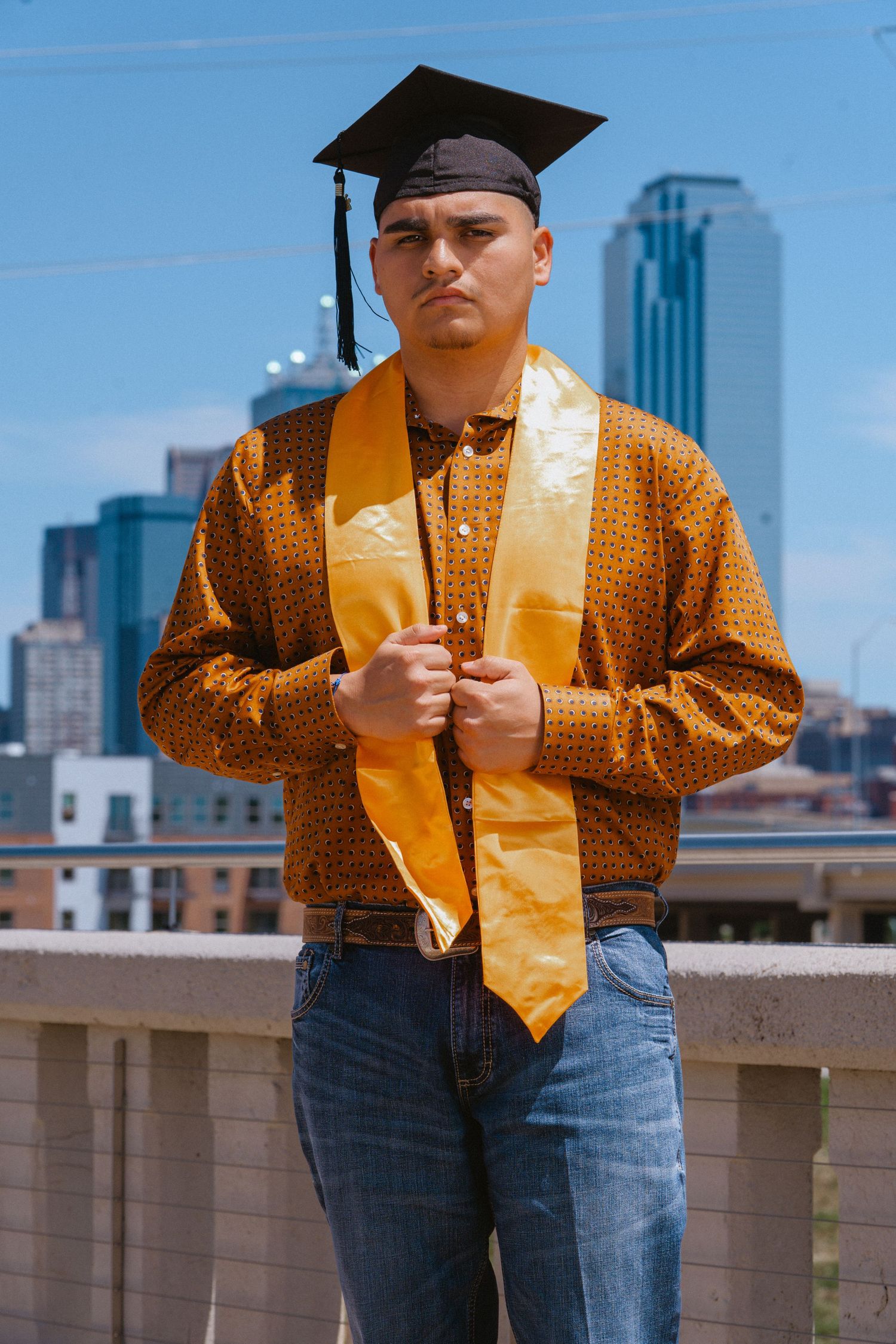 Graduate in gold stole and cap poses confidently with city skyline behind him.