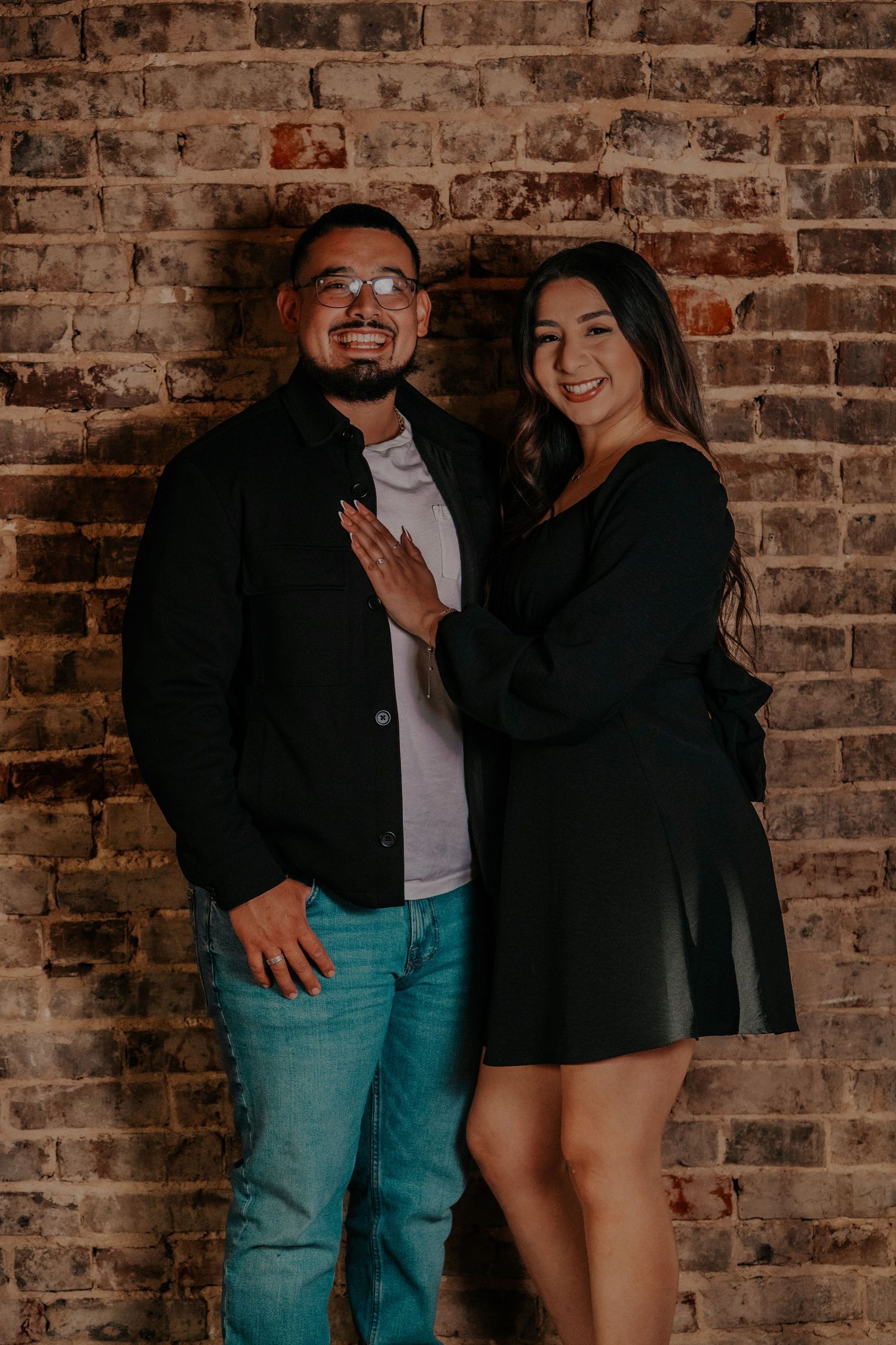 Couple laughing and posing against rustic brick wall in casual outfits during photo session.