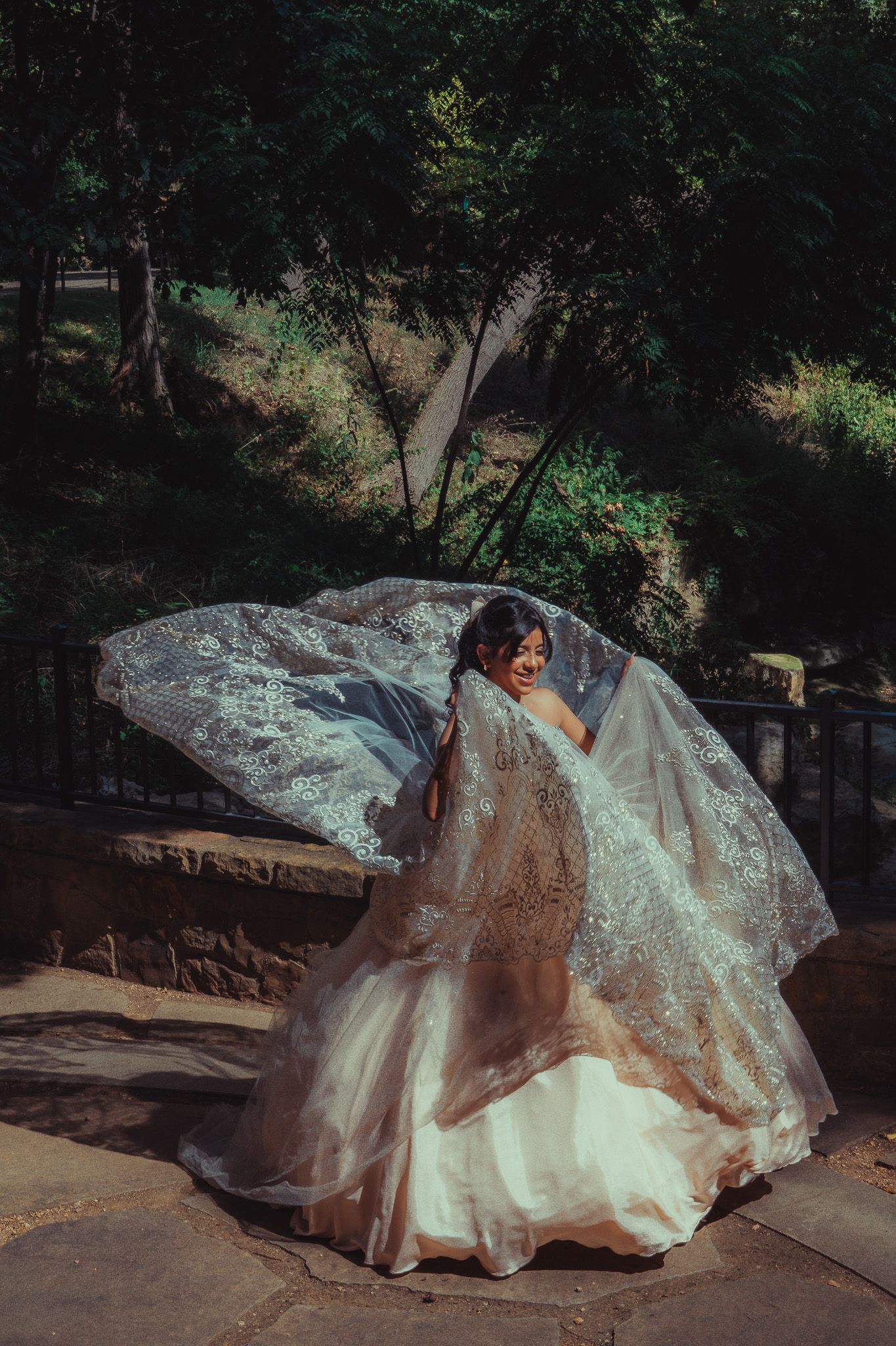 Bride in lace wedding gown with flowing veil billowing in wind, posing outdoors in sunlit garden setting.