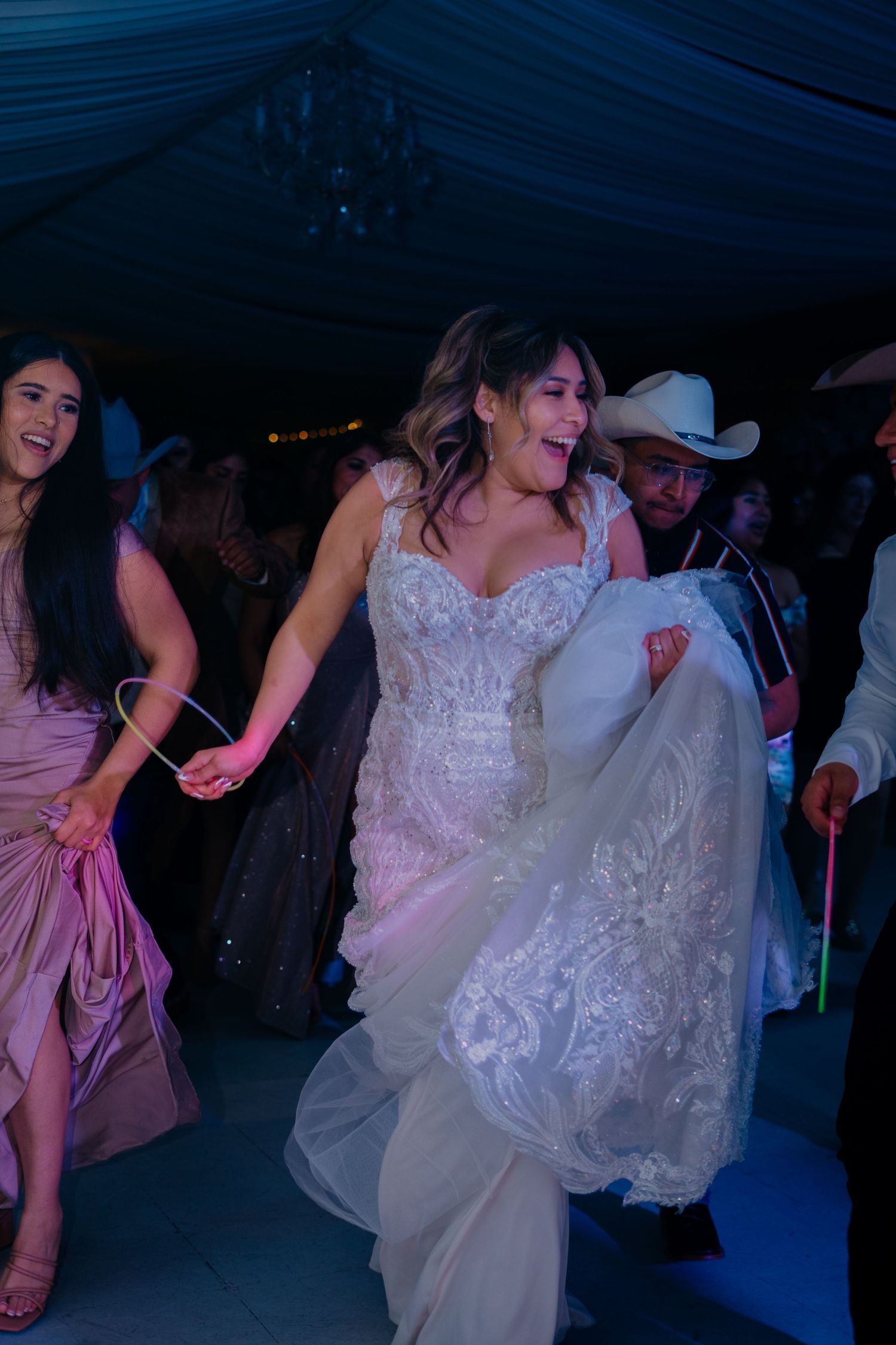 A bride in a white lace wedding dress dances joyfully at a reception under blue lighting with other guests.