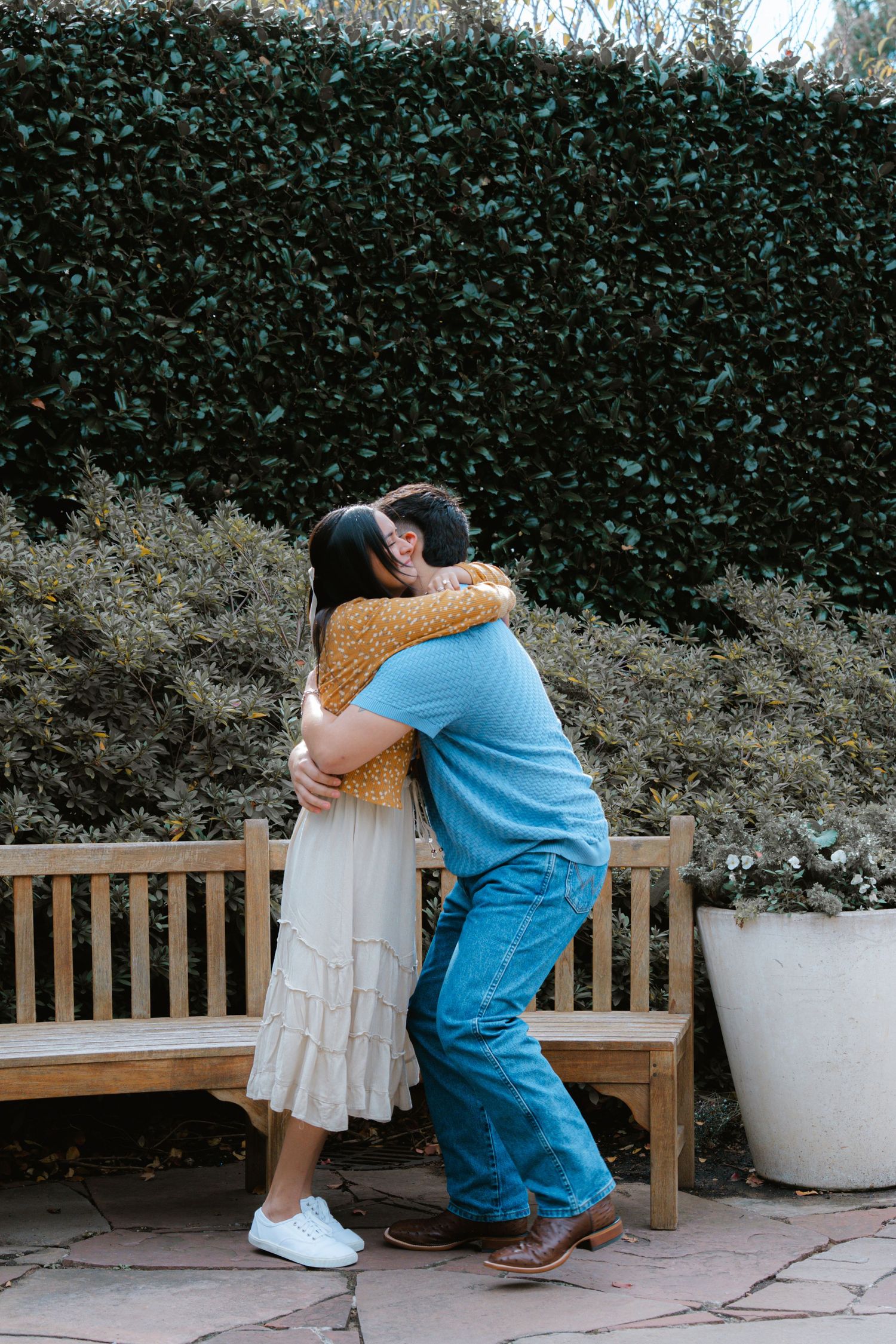 A couple shares an intimate moment on a wooden bench against a hedge wall in an outdoor setting.