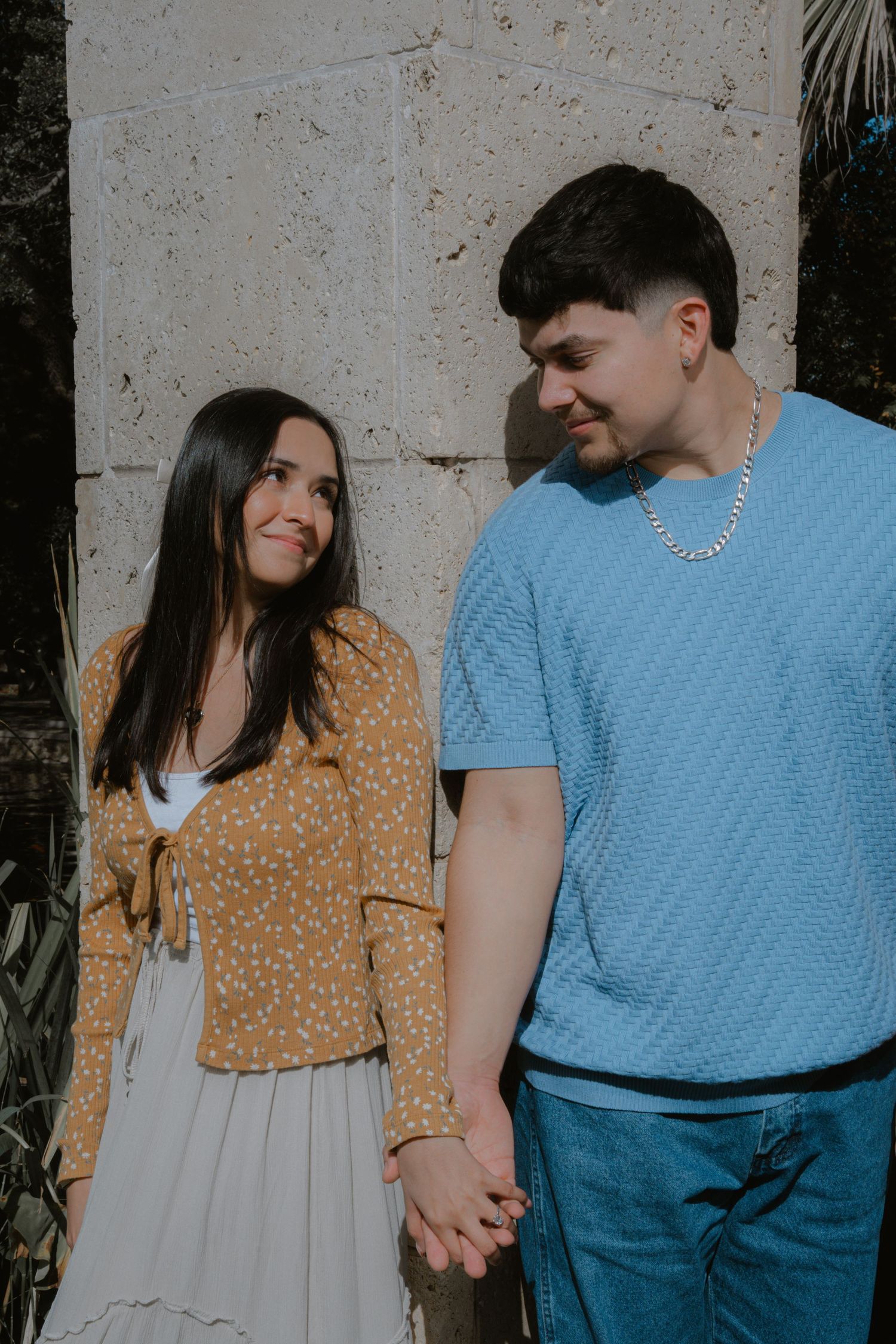 A couple in casual attire holding hands against a stone wall backdrop.