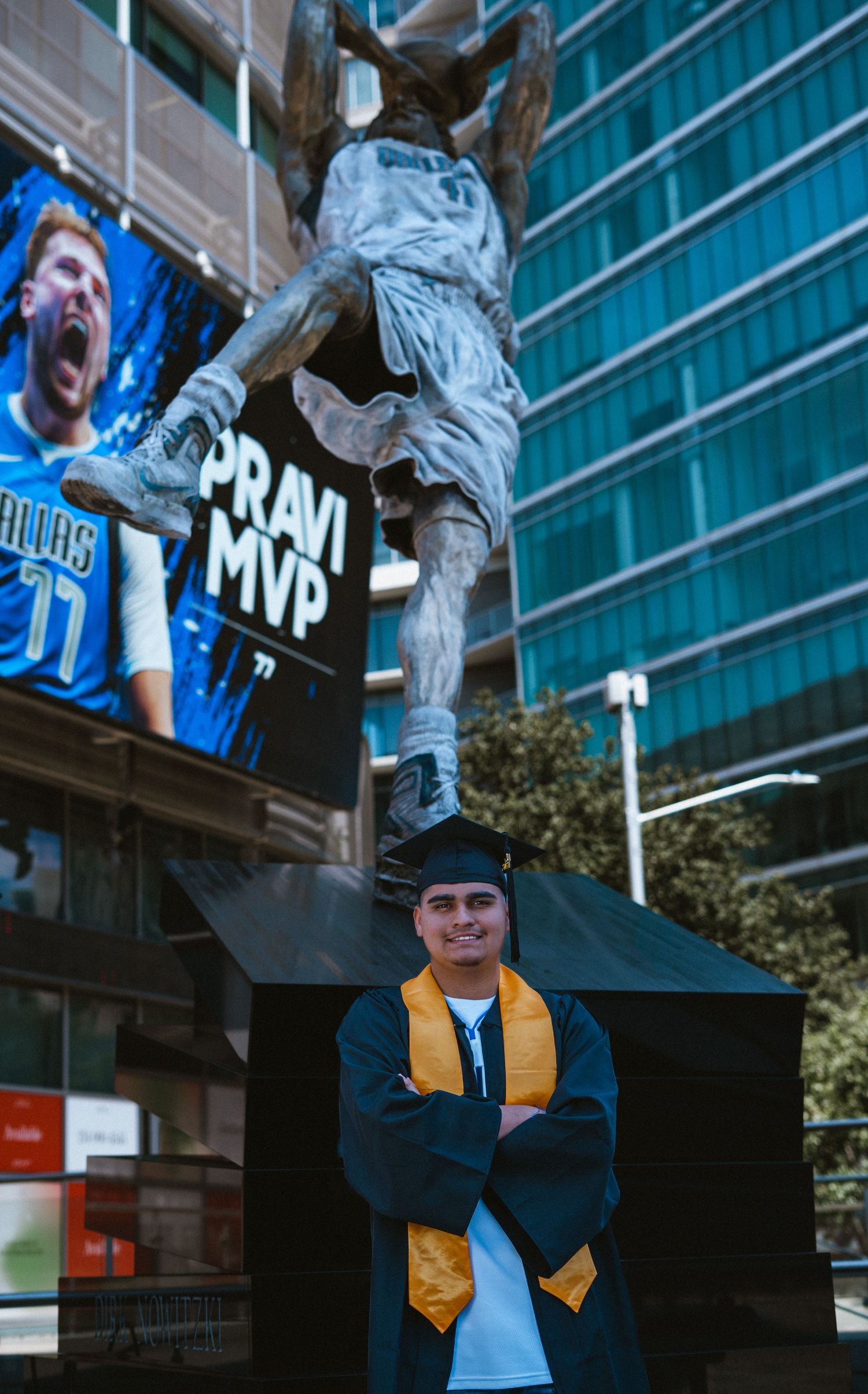 Graduate in cap and gown poses arms crossed under a basketball statue with MVP billboard backdrop.