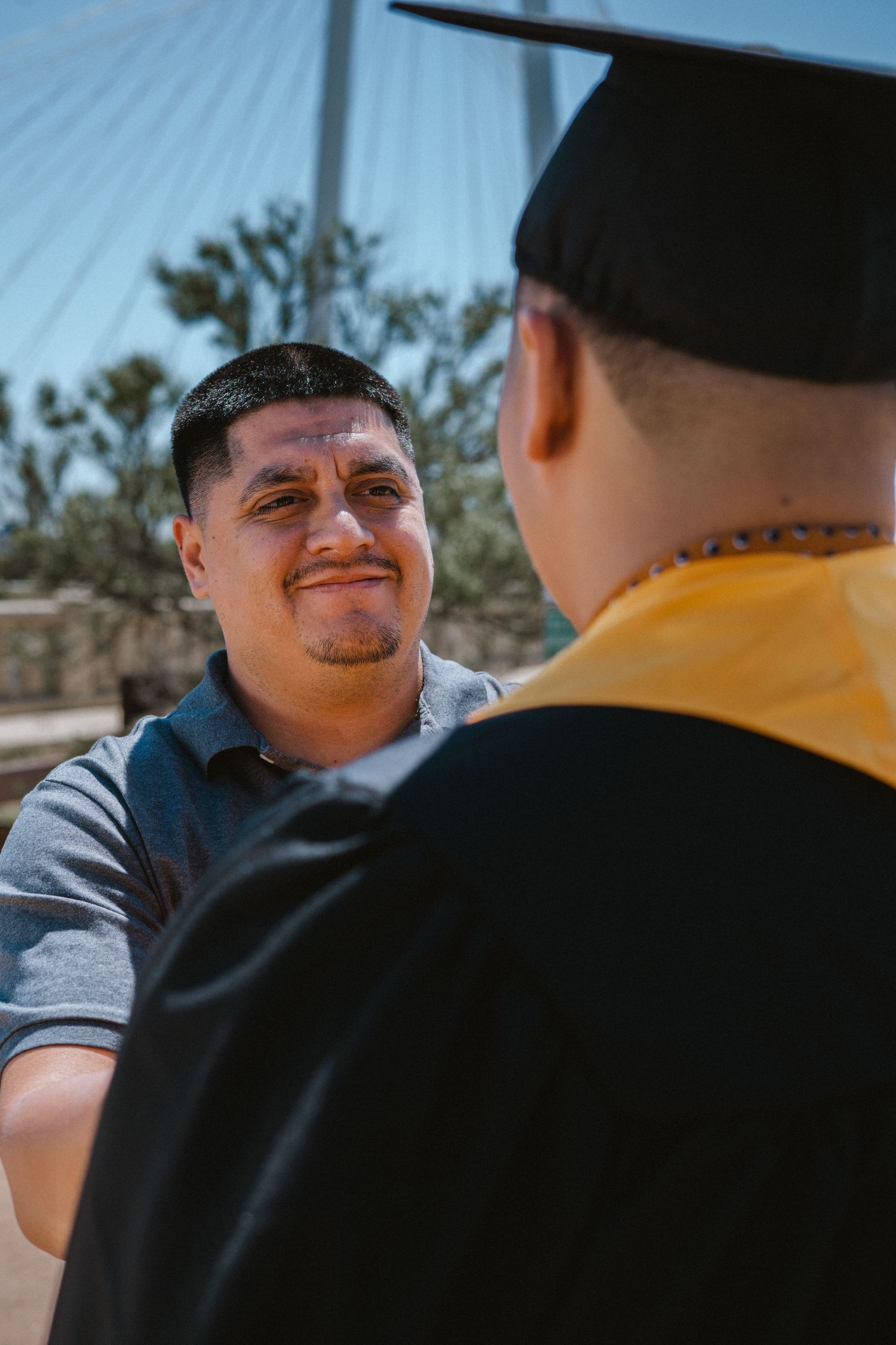 A smiling young man congratulates a graduate in black cap and yellow stole outdoors near a bridge.