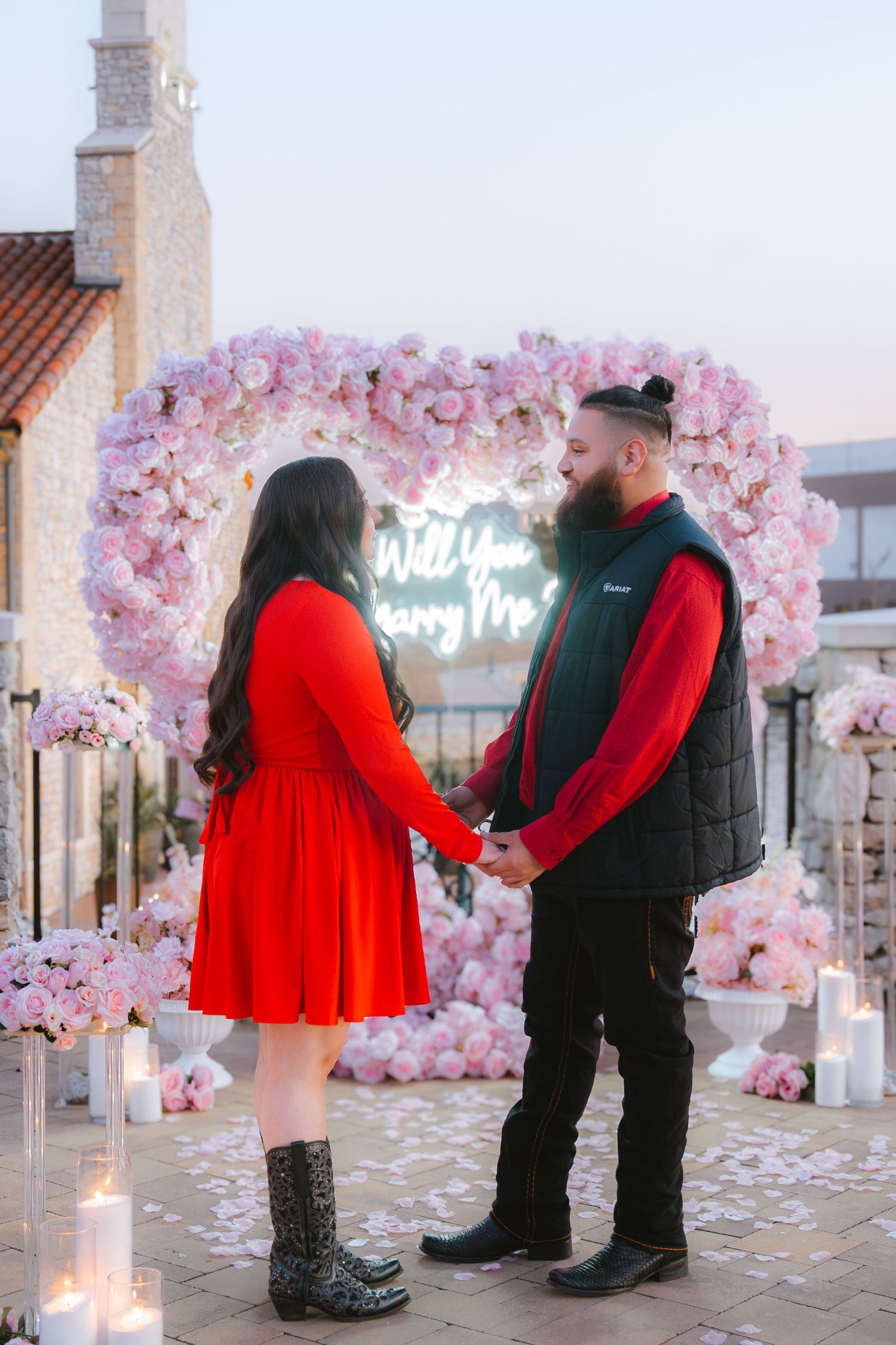 Couple holds hands during romantic marriage proposal with pink floral heart arch and Will You Marry Me neon sign.