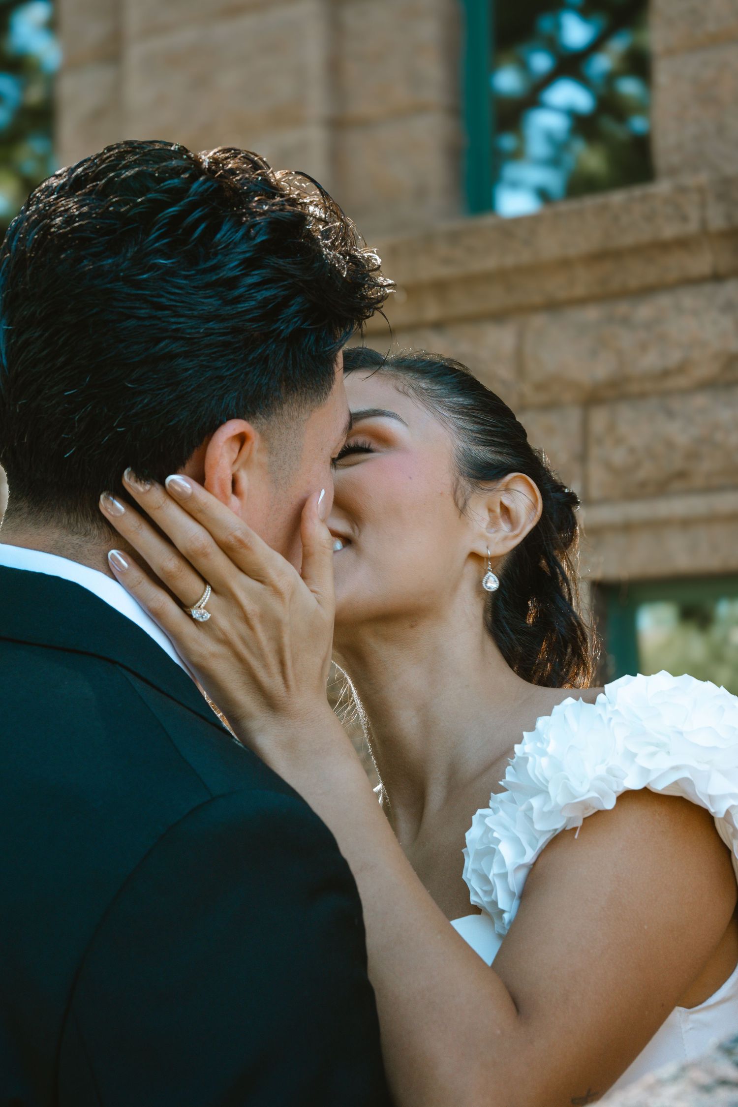 A bride and groom share a romantic kiss, with the bride wearing a white ruffled dress and diamond ring.
