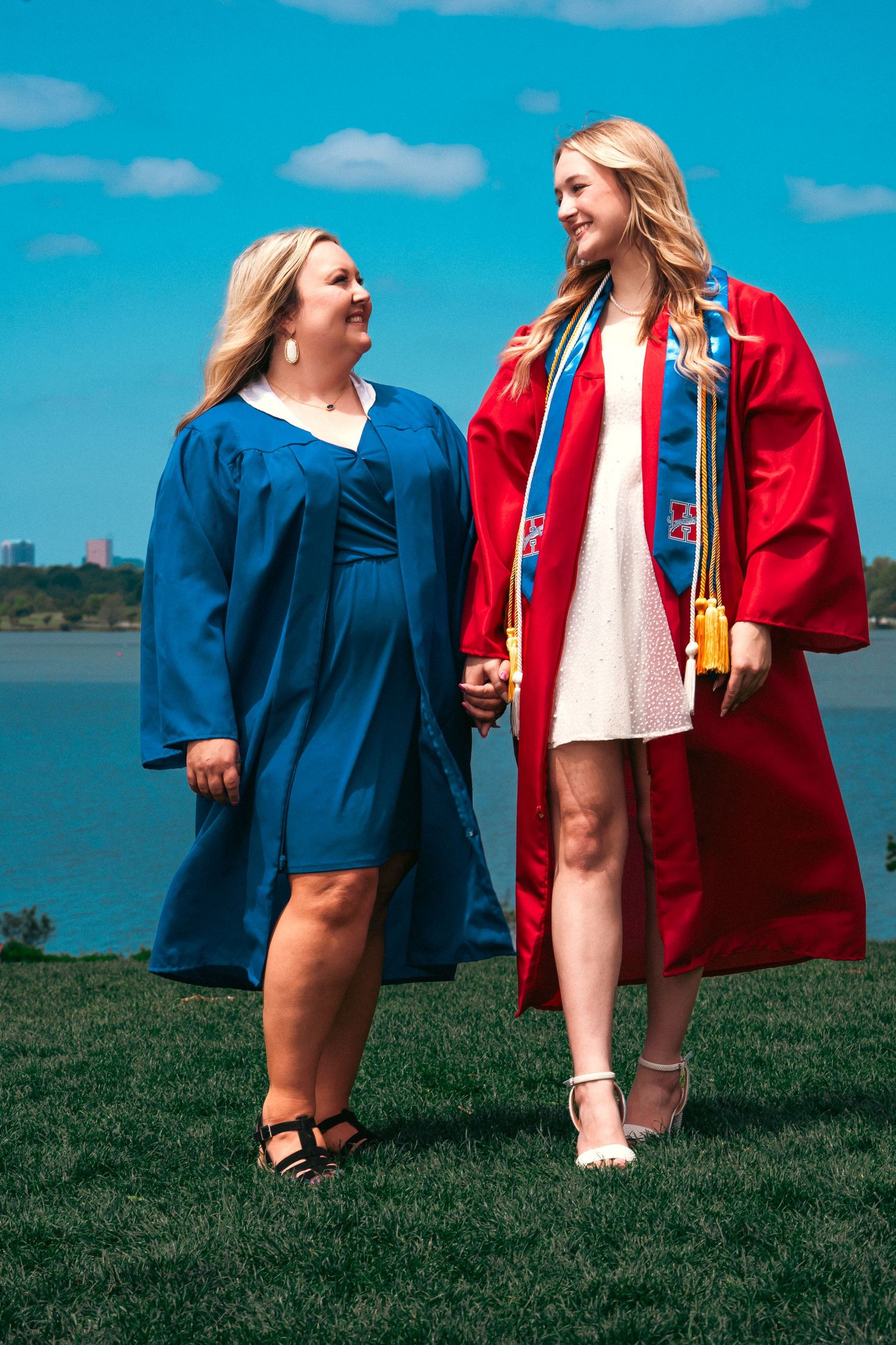 Two graduates in blue and red gowns celebrate on a grassy hill overlooking a lake on a sunny day.