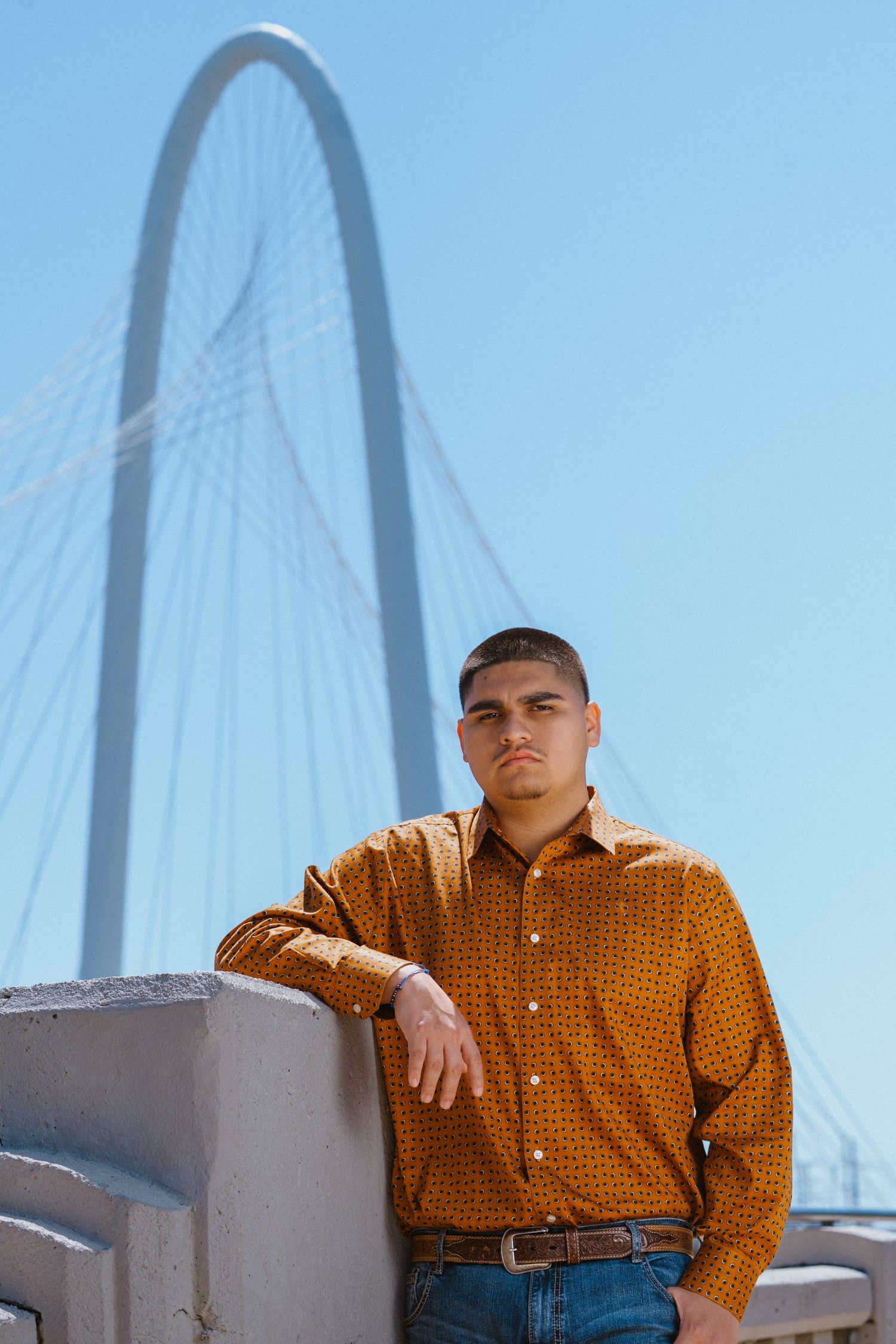 Young man in orange patterned shirt leaning on concrete railing with Margaret Hunt Hill Bridge behind him.