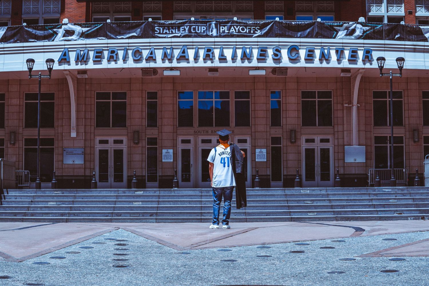 Fan stands outside American Airlines Center during Stanley Cup Playoffs, wearing a sports jersey on a brick plaza.