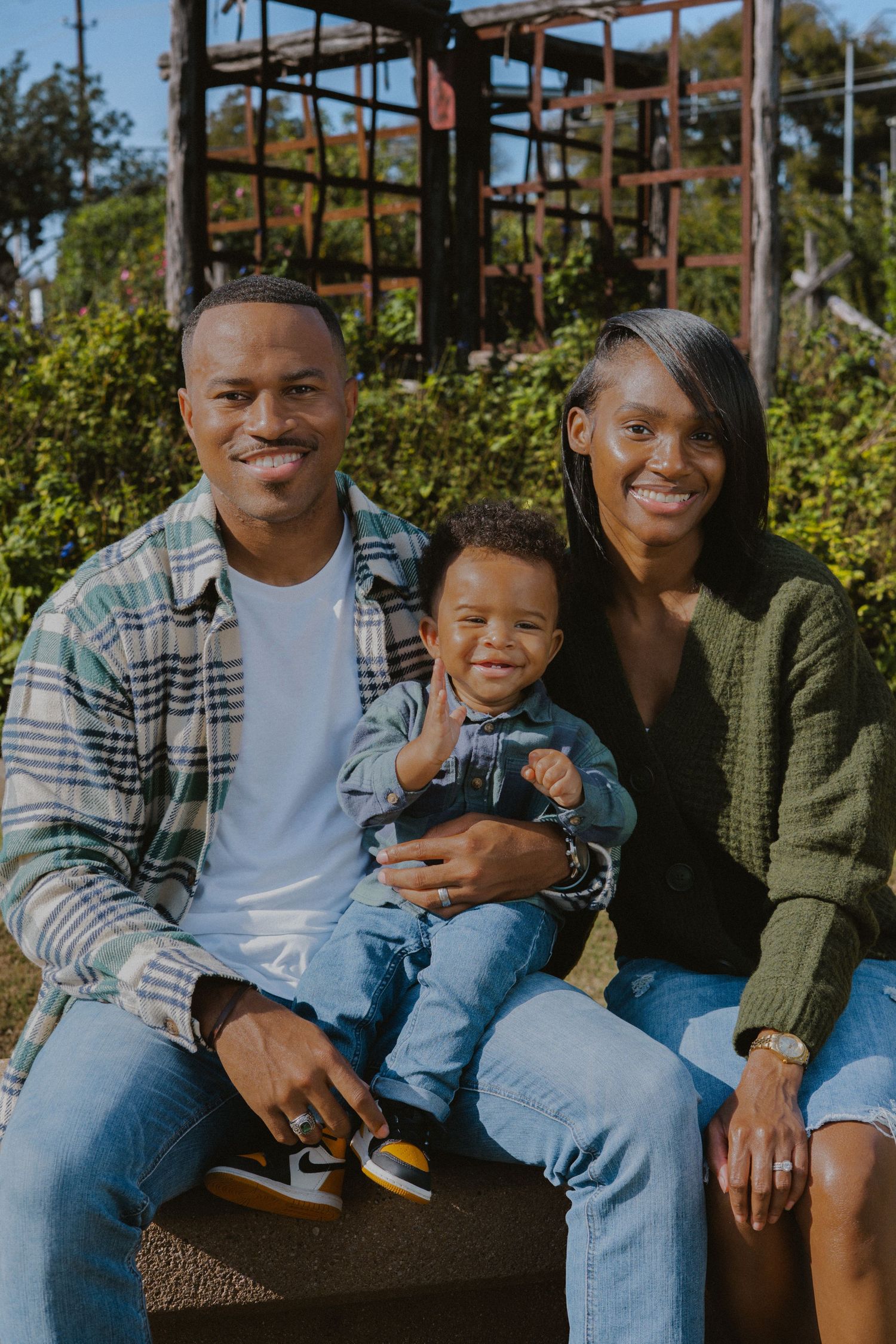Smiling Black family portrait outdoors, parents in casual attire holding adorable baby in denim outfit.
