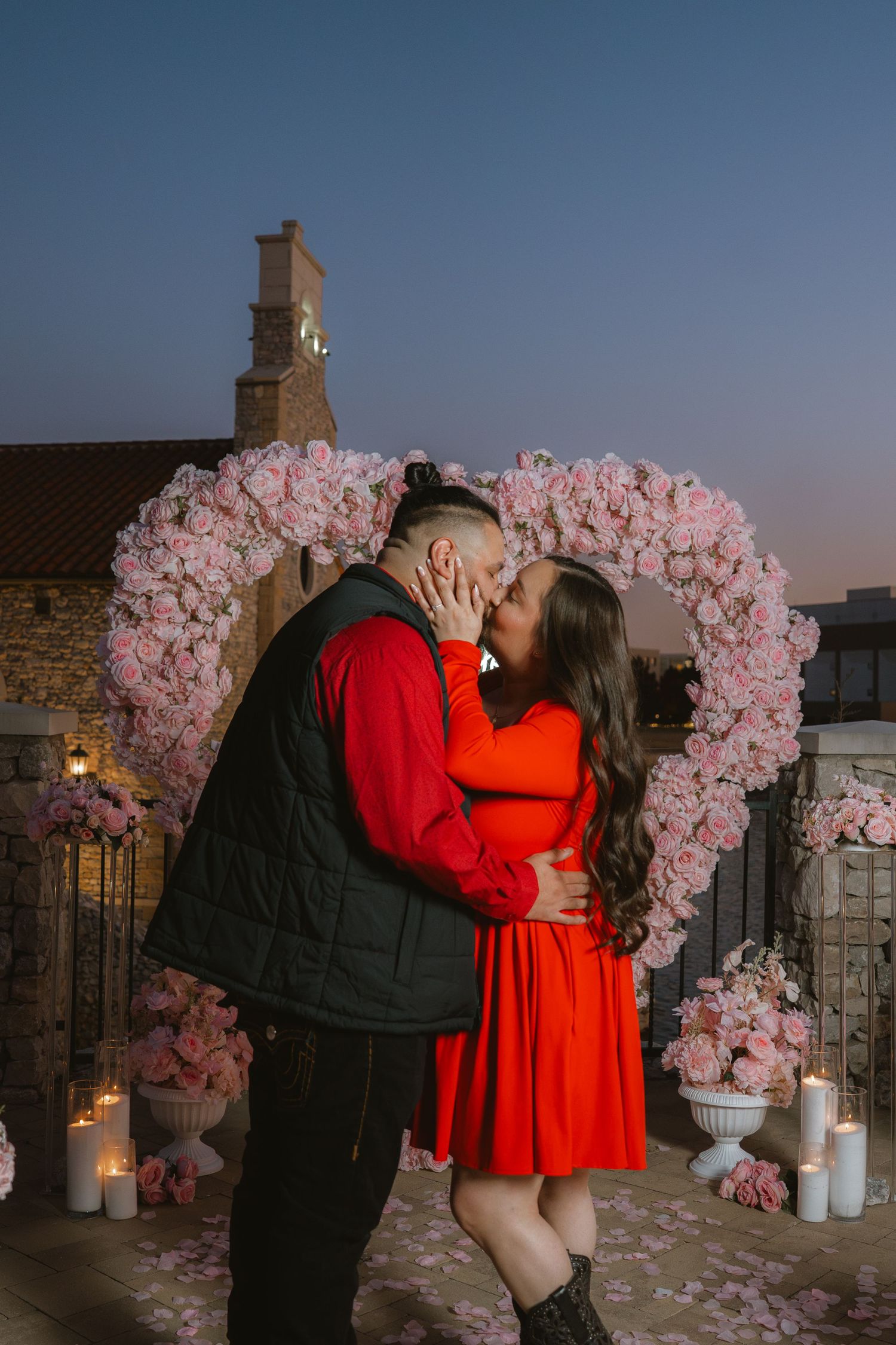 Couple kissing under pink floral heart arch with candles at dusk during romantic proposal.