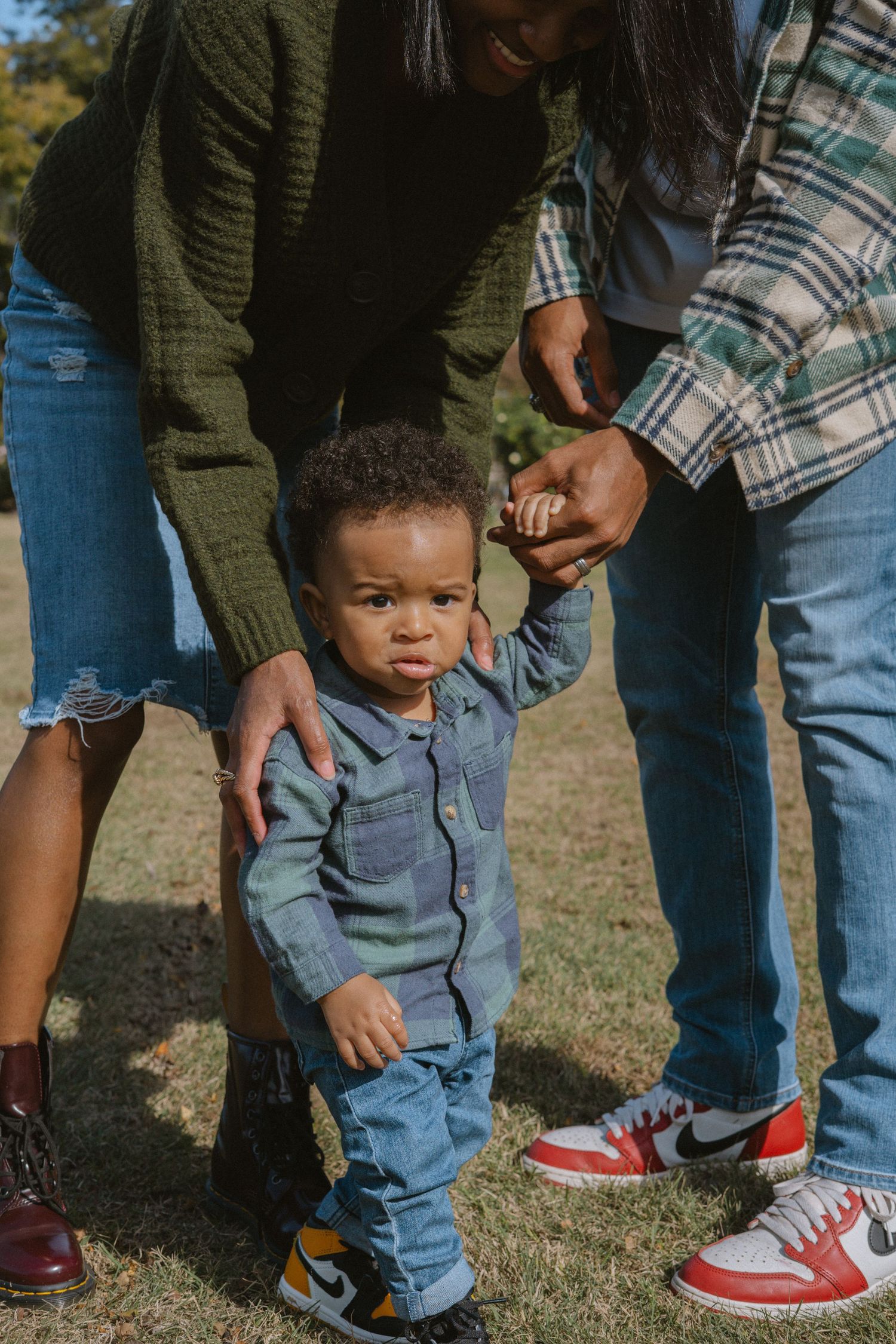 A toddler in denim outfit takes first steps outdoors, supported by two adults in casual clothes on green grass.