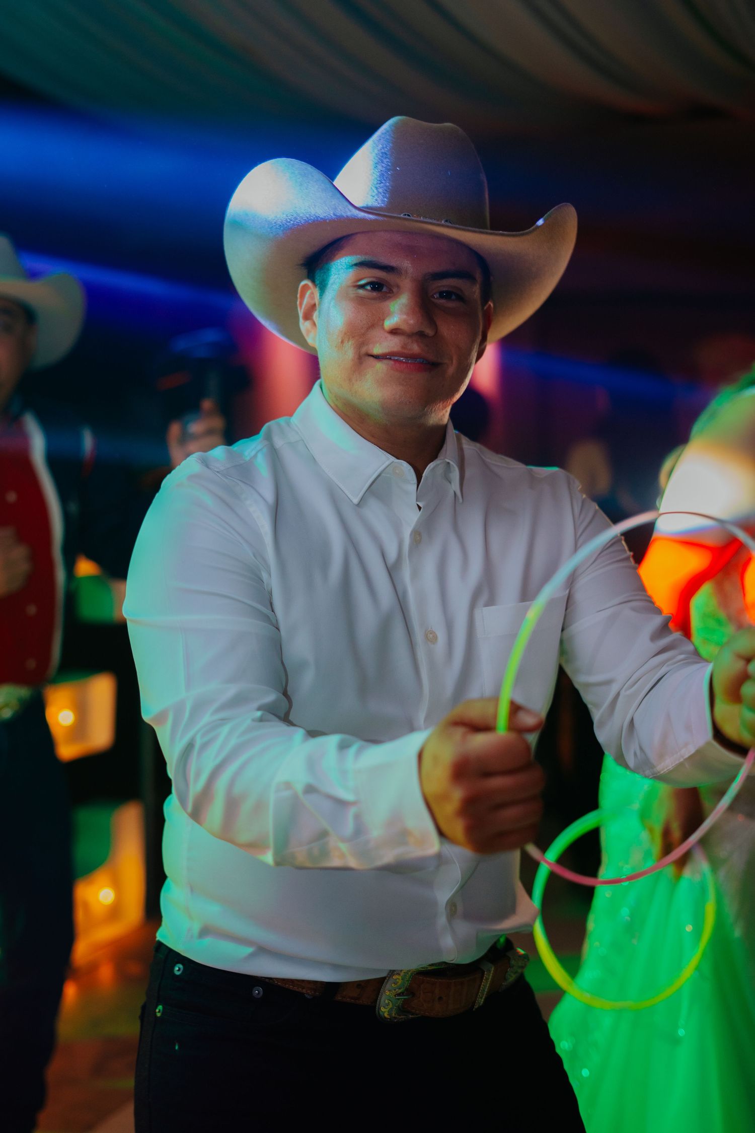A person in a white cowboy hat and shirt dances under colorful nightclub lighting with neon green accents.