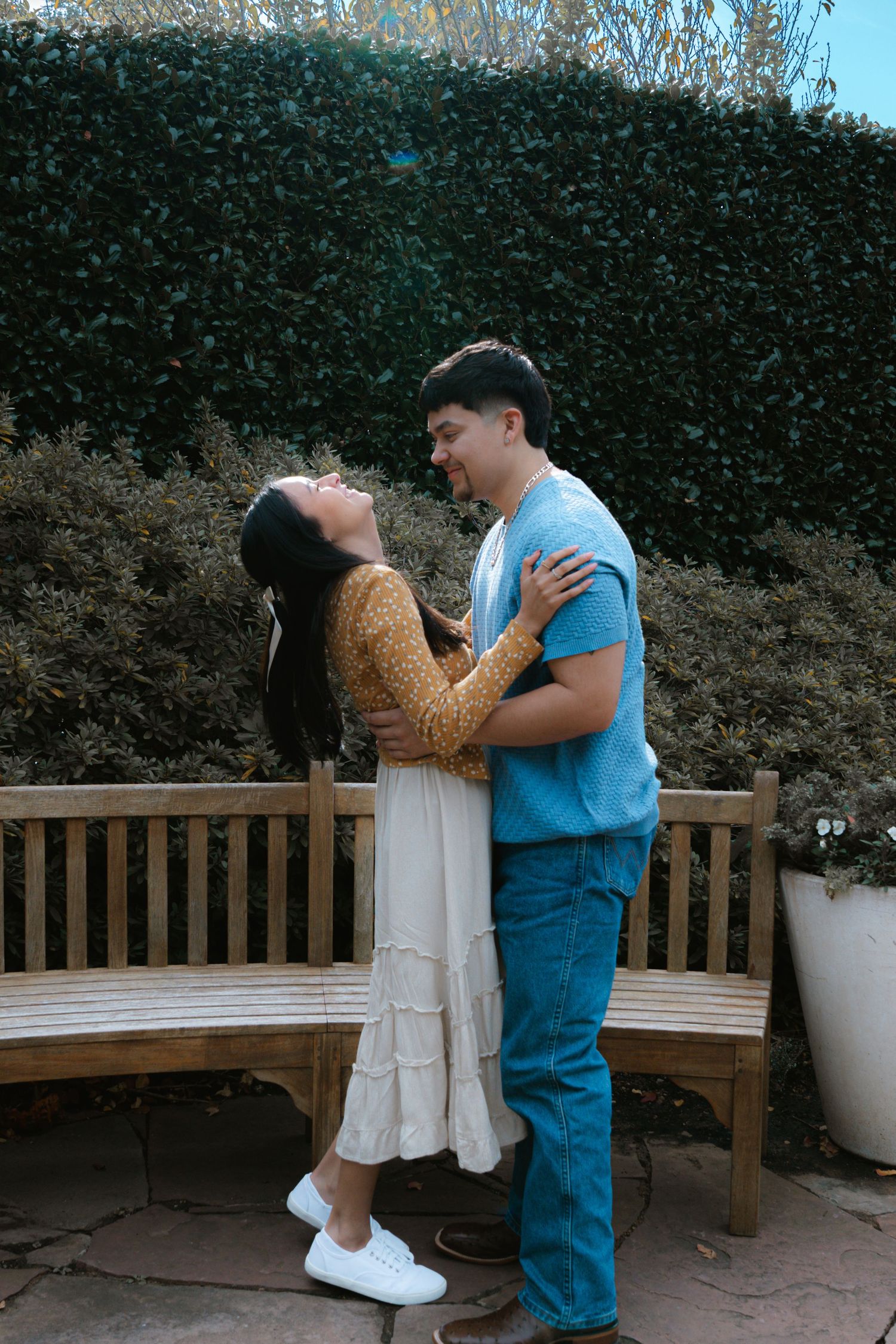 A romantic proposal scene unfolds on a wooden bench surrounded by greenery.