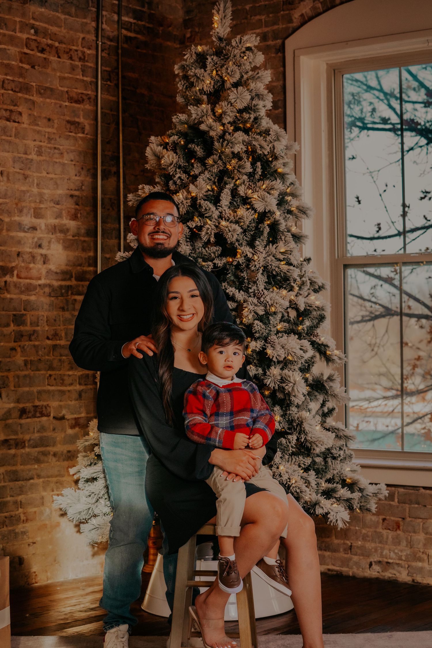 A family shares joyful moments together in front of a decorated Christmas tree in a rustic brick-walled room.