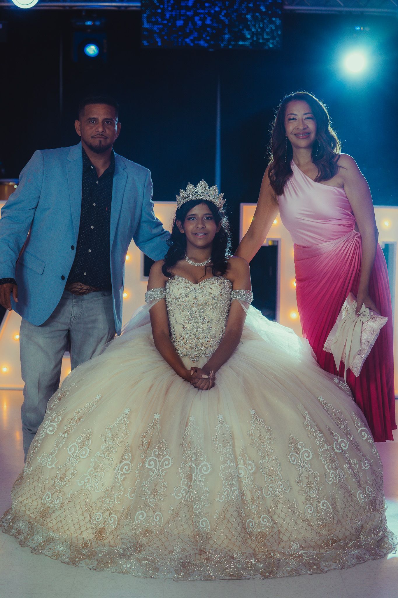 Young woman in ornate white quinceañera gown and tiara poses with two adults at elegant celebration event.
