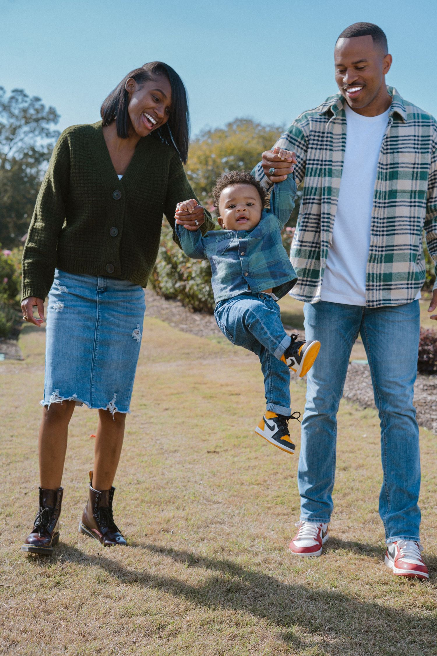 A joyful Black family swings their toddler between them outdoors, all wearing jeans and colorful sneakers on a sunny day.
