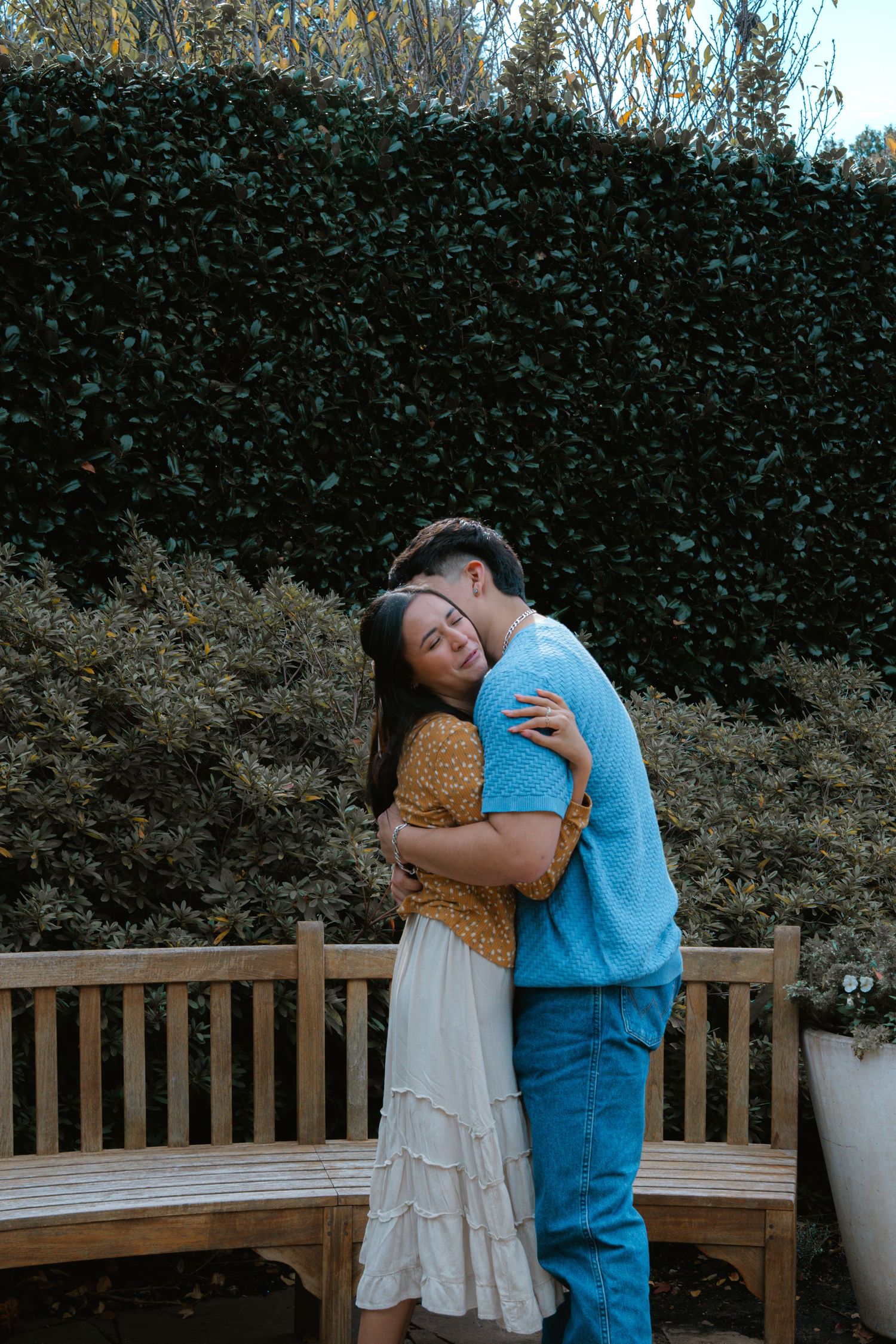 Two people embrace emotionally on a wooden bench surrounded by greenery and white planters.