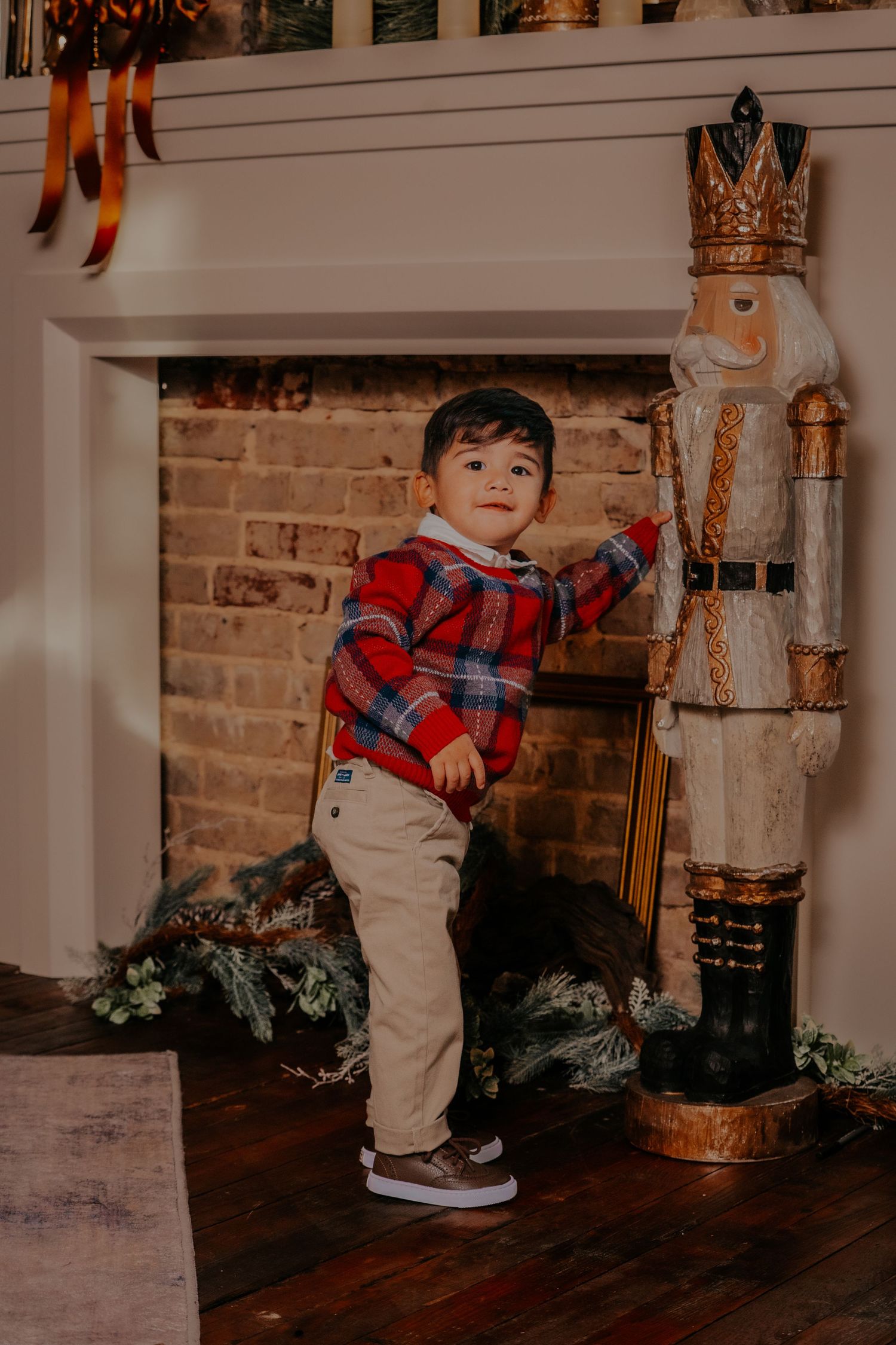 A toddler in a red plaid shirt and khaki pants stands by a rustic brick wall with holiday decorations.