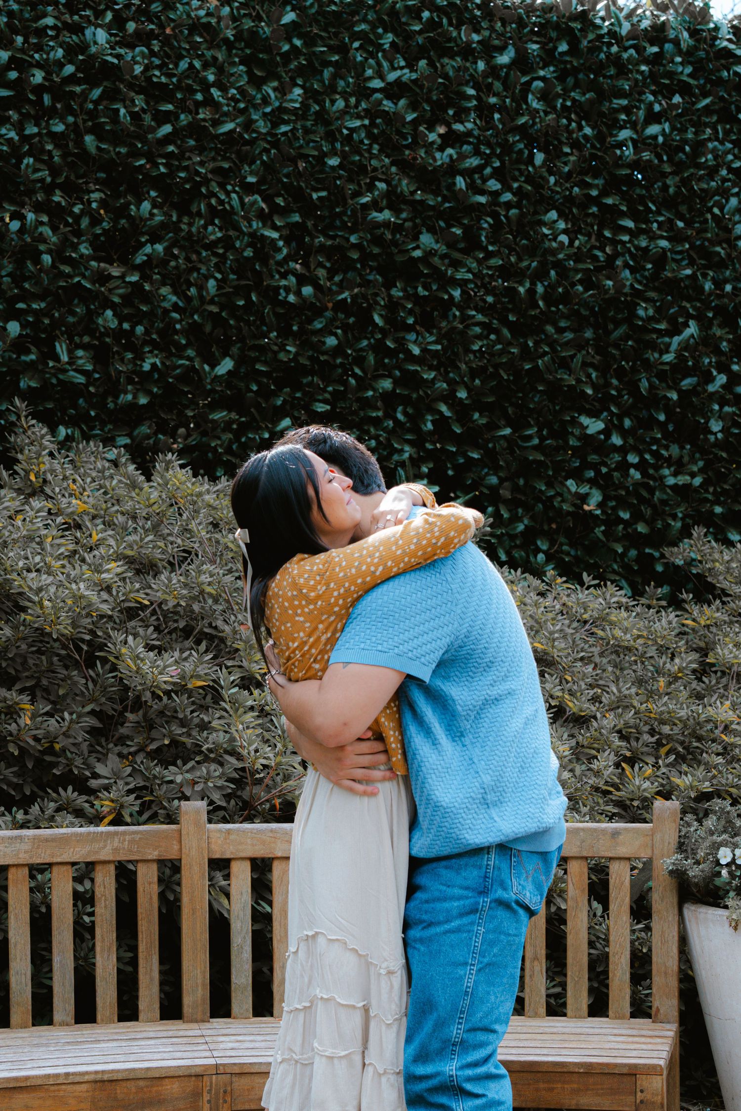 Two people embrace while sitting on a garden bench surrounded by greenery.