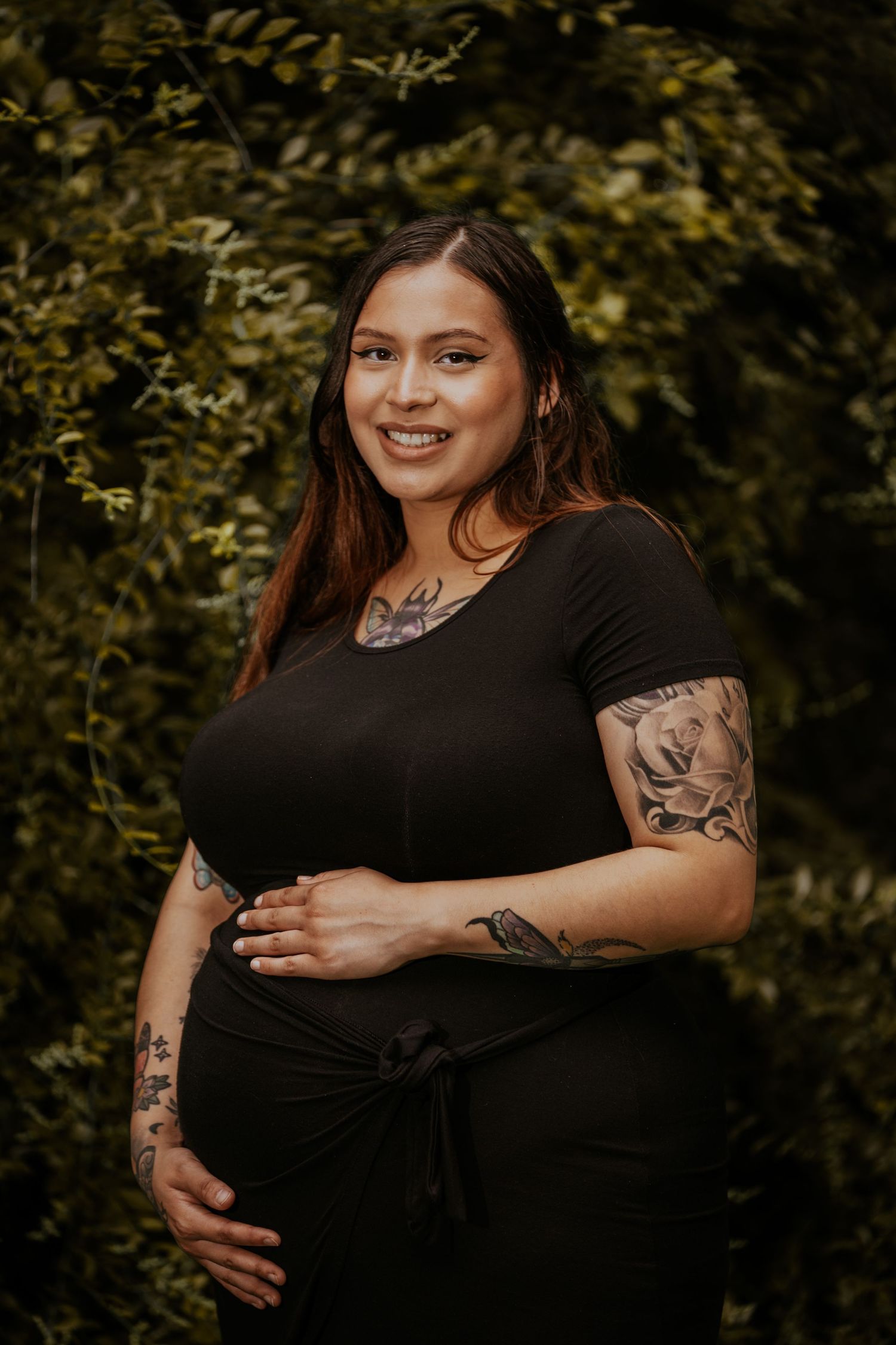 Maternity photoshoot in a garden setting with black dress and visible arm tattoos against lush green foliage.