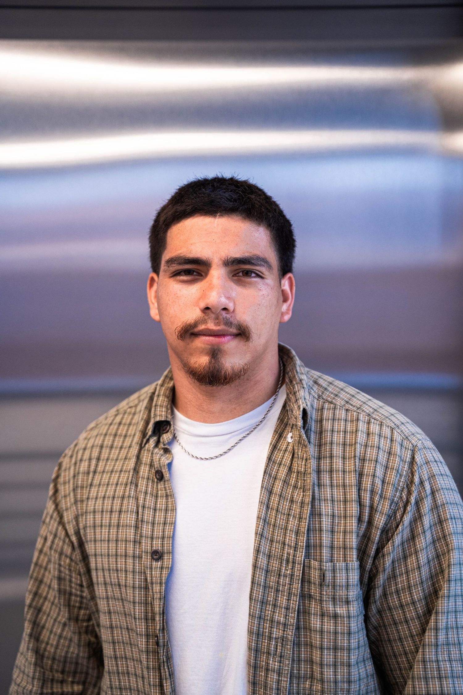 Young man with goatee wearing plaid shirt and chain necklace standing in front of metallic background.