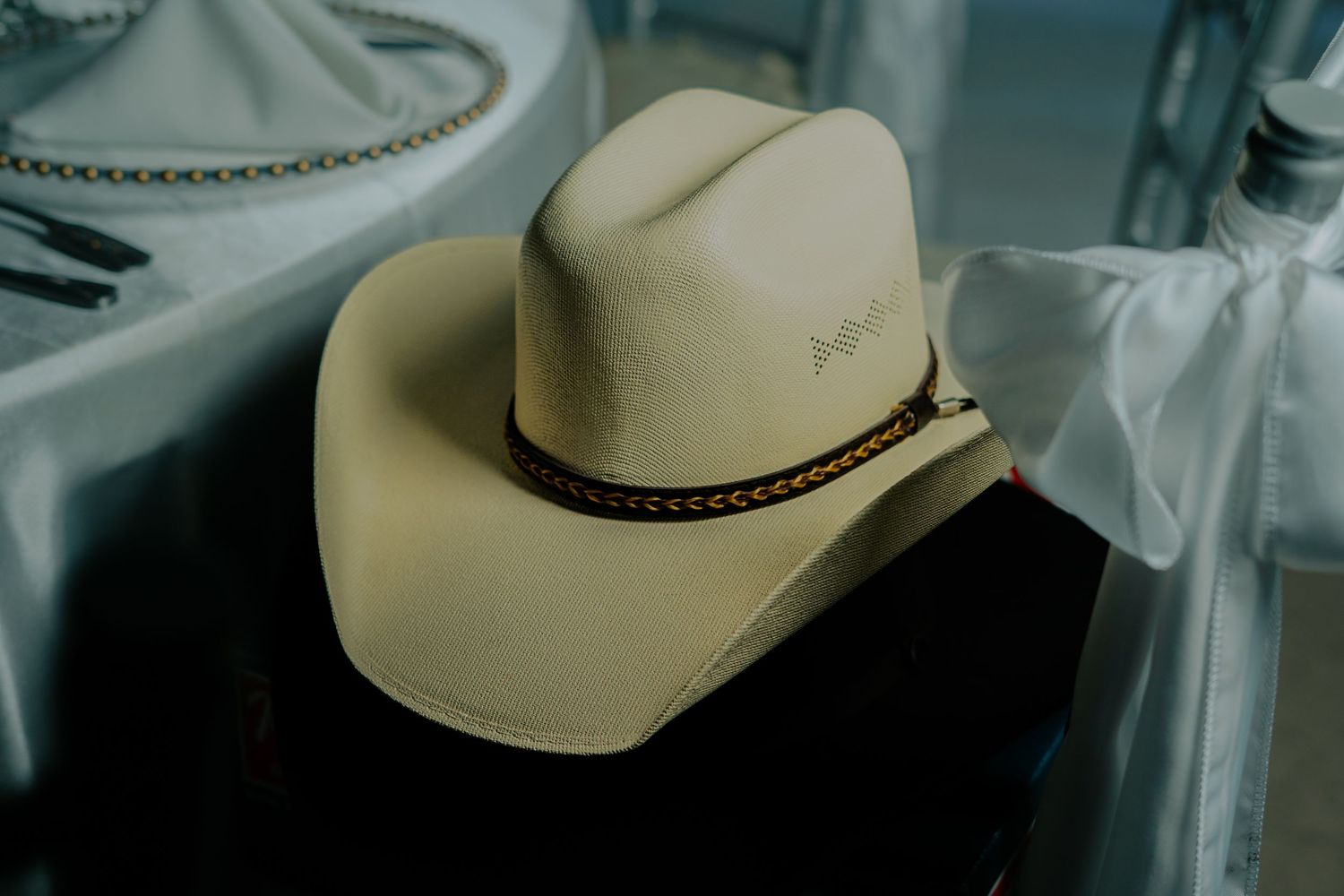A beige cowboy hat with braided leather band sits on a reflective surface in moody lighting.