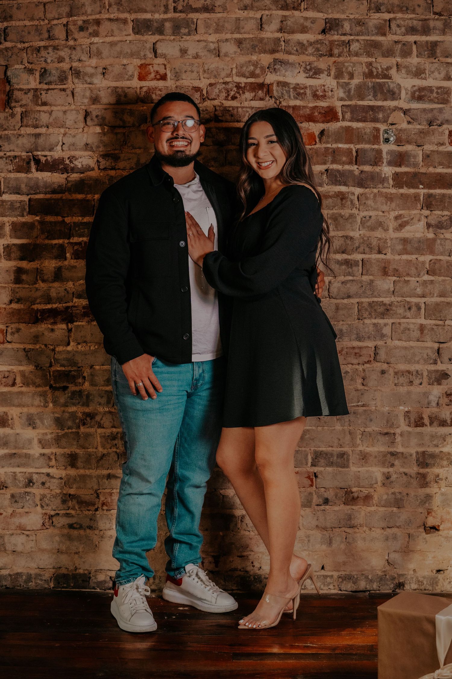 A couple poses playfully against a rustic brick wall in a casual photoshoot setting.