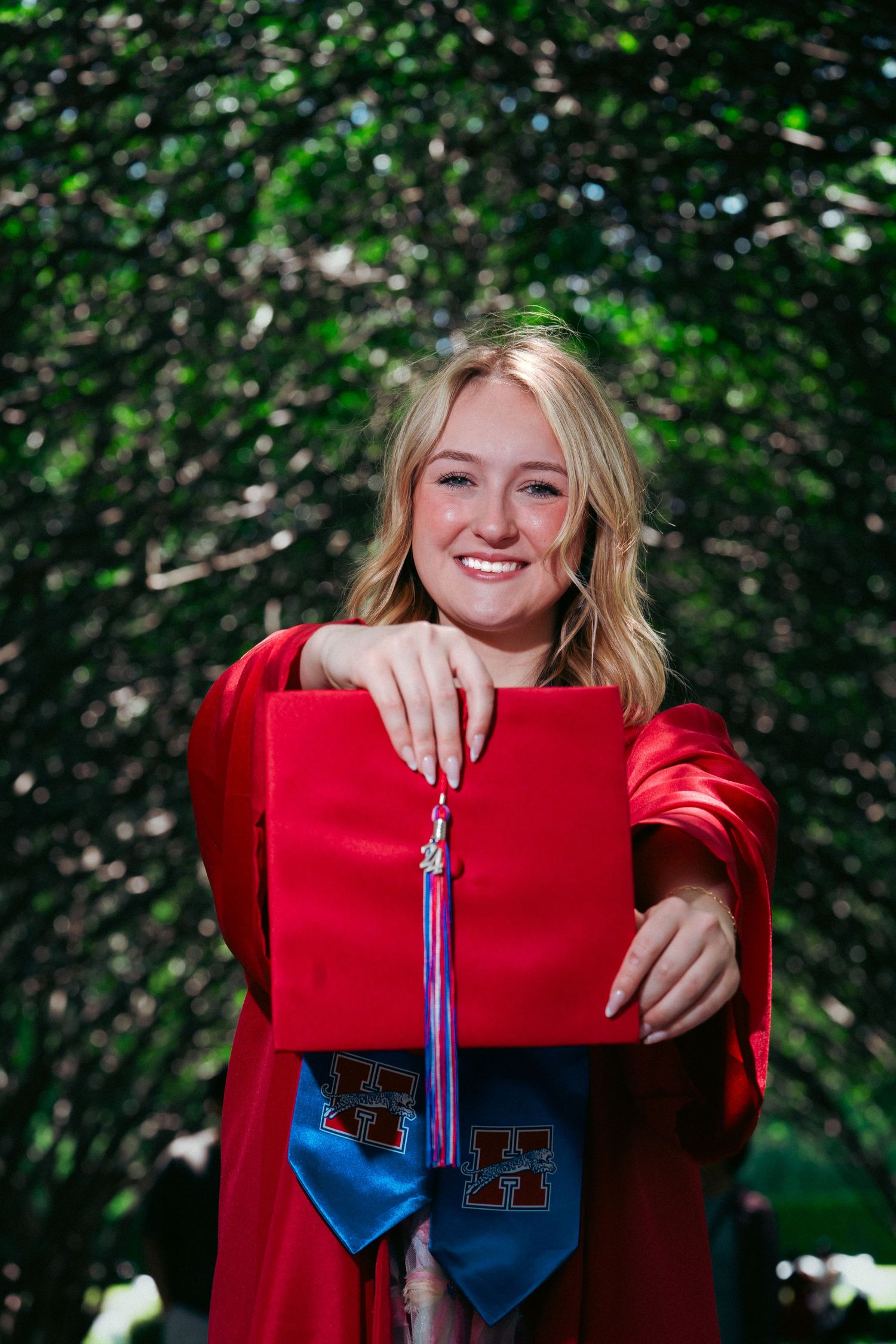 Graduate in red cap and gown holds diploma with blue tassel against green foliage background.