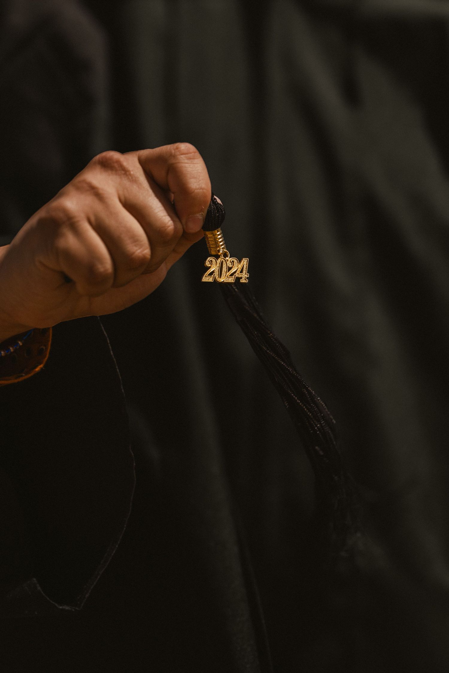 Hand holding a black graduation tassel with a gold 2024 charm, wearing a black graduation gown.