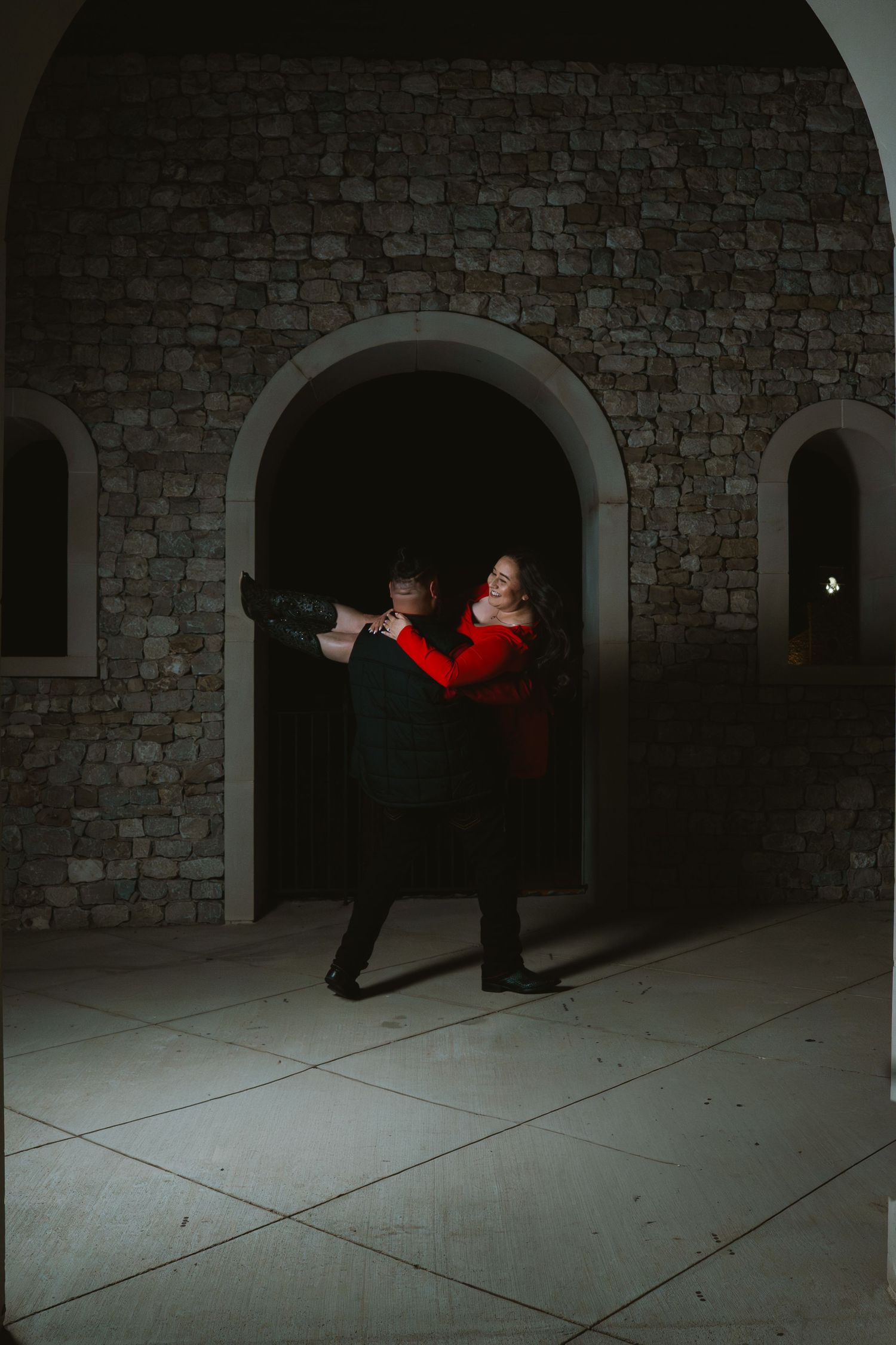 Couple dancing romantically in front of a stone arch doorway in dramatic lighting, woman wearing red dress.