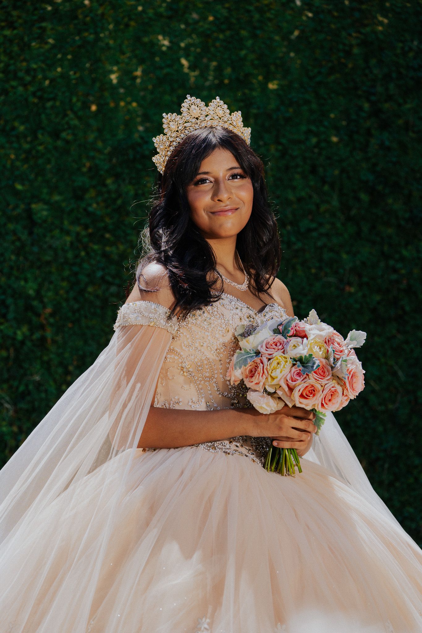 Young woman in a blush ballgown and gold crown holds a pastel rose bouquet, posing outdoors for her quinceañera.